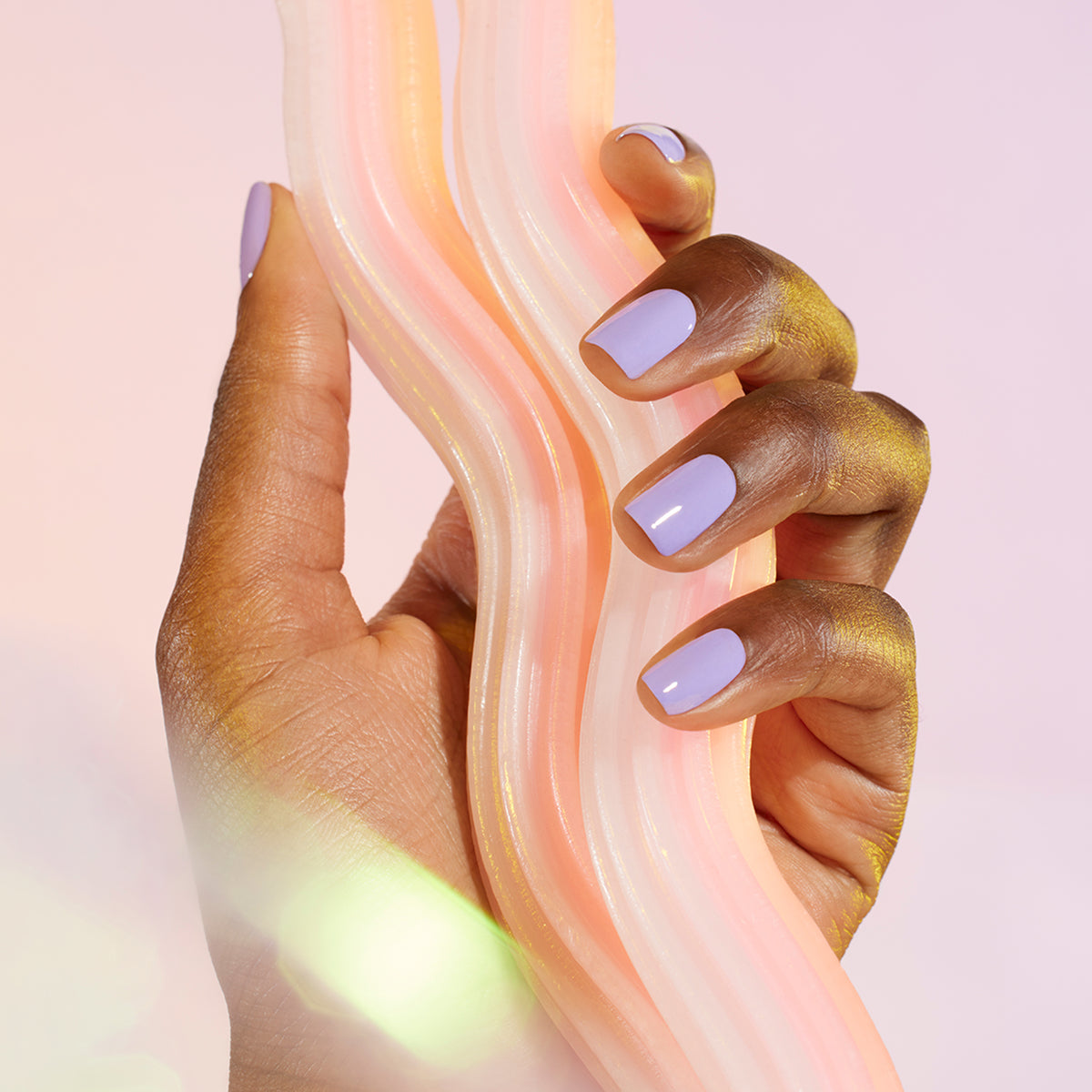 Une main avec le vernis à ongles violet pastel Wisteria de Manucurist tient un objet ondulé et translucide aux sous-tons bleu froid sur un fond rose doux.