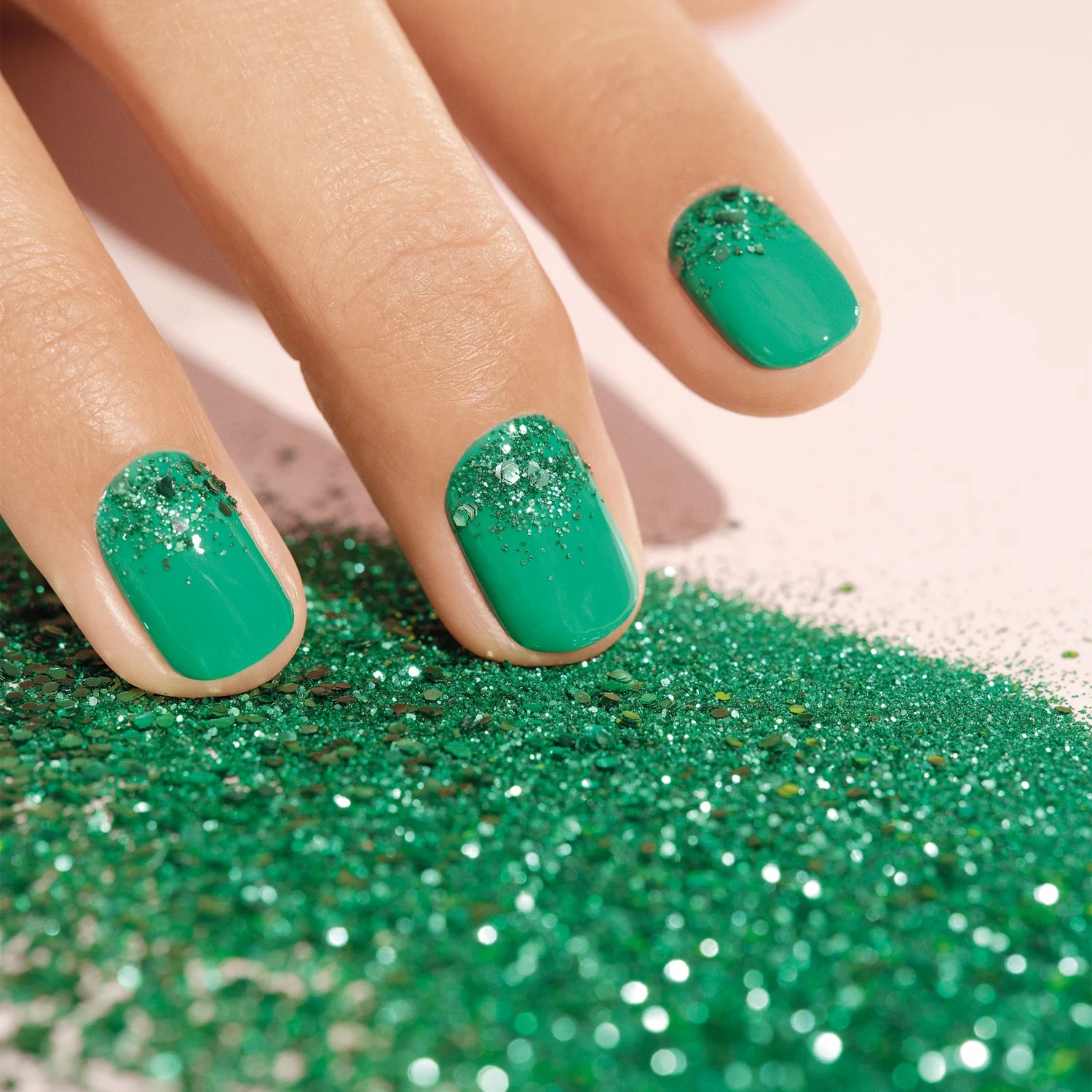 A hand with green nail polish and silver glitter on the tips of the nails rests on a surface covered in green glitter.