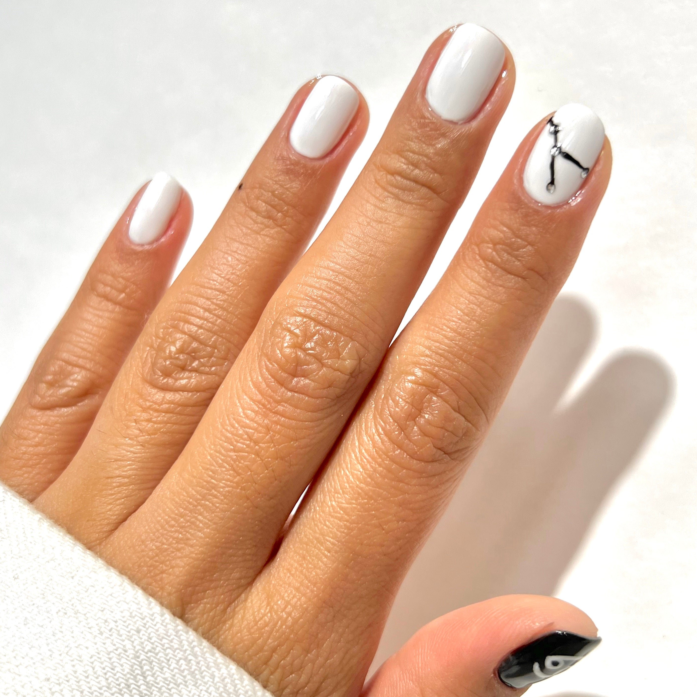 A hand with short, neatly manicured nails painted white, except the thumb which is painted black with a white eye design. The ring fingernail features a delicate black line art design.