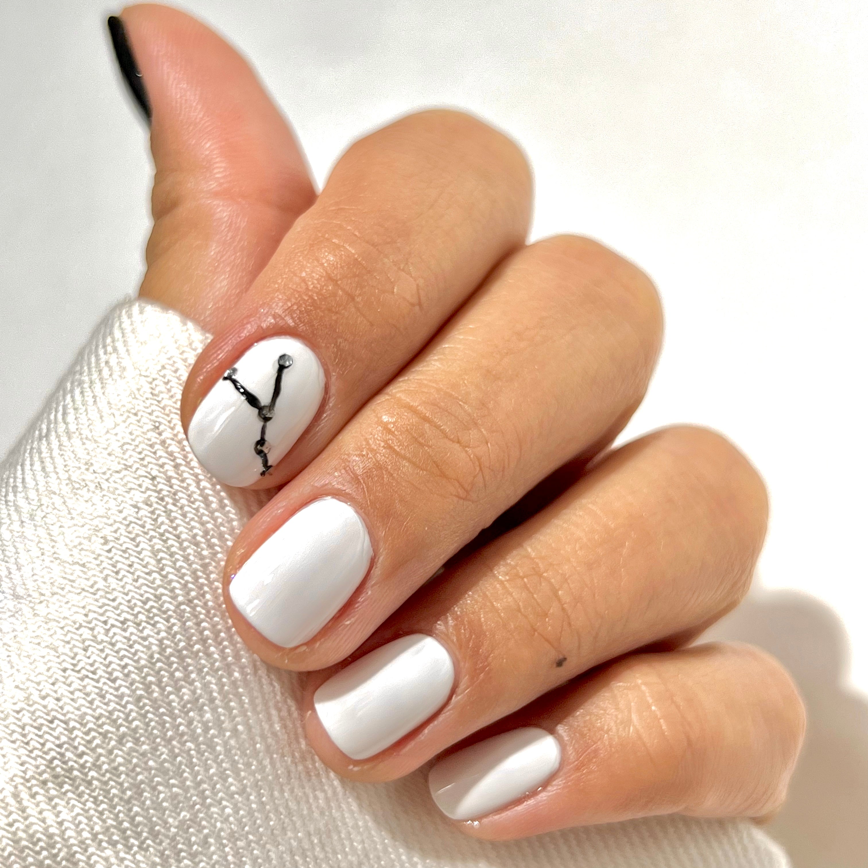 A hand with short, square-shaped nails painted white; the ring fingernail features a simple black constellation-like line design. The person is wearing a white, long-sleeve top that covers part of their hand.