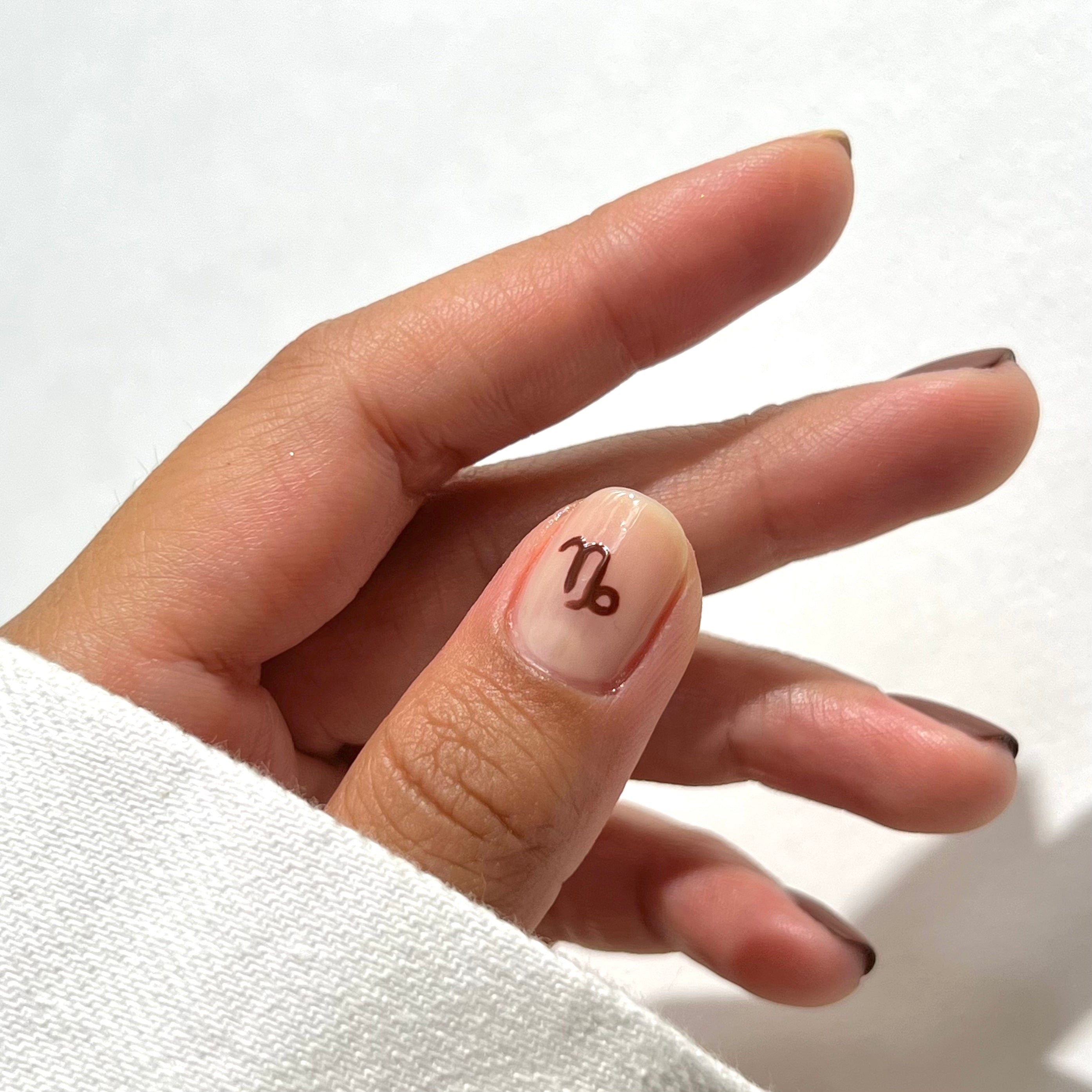 A hand with neatly manicured nails, each tipped with a thin brown line, and the thumb nail featuring a brown Capricorn zodiac symbol. The hand is partially covered by a white sleeve, set against a plain white background.