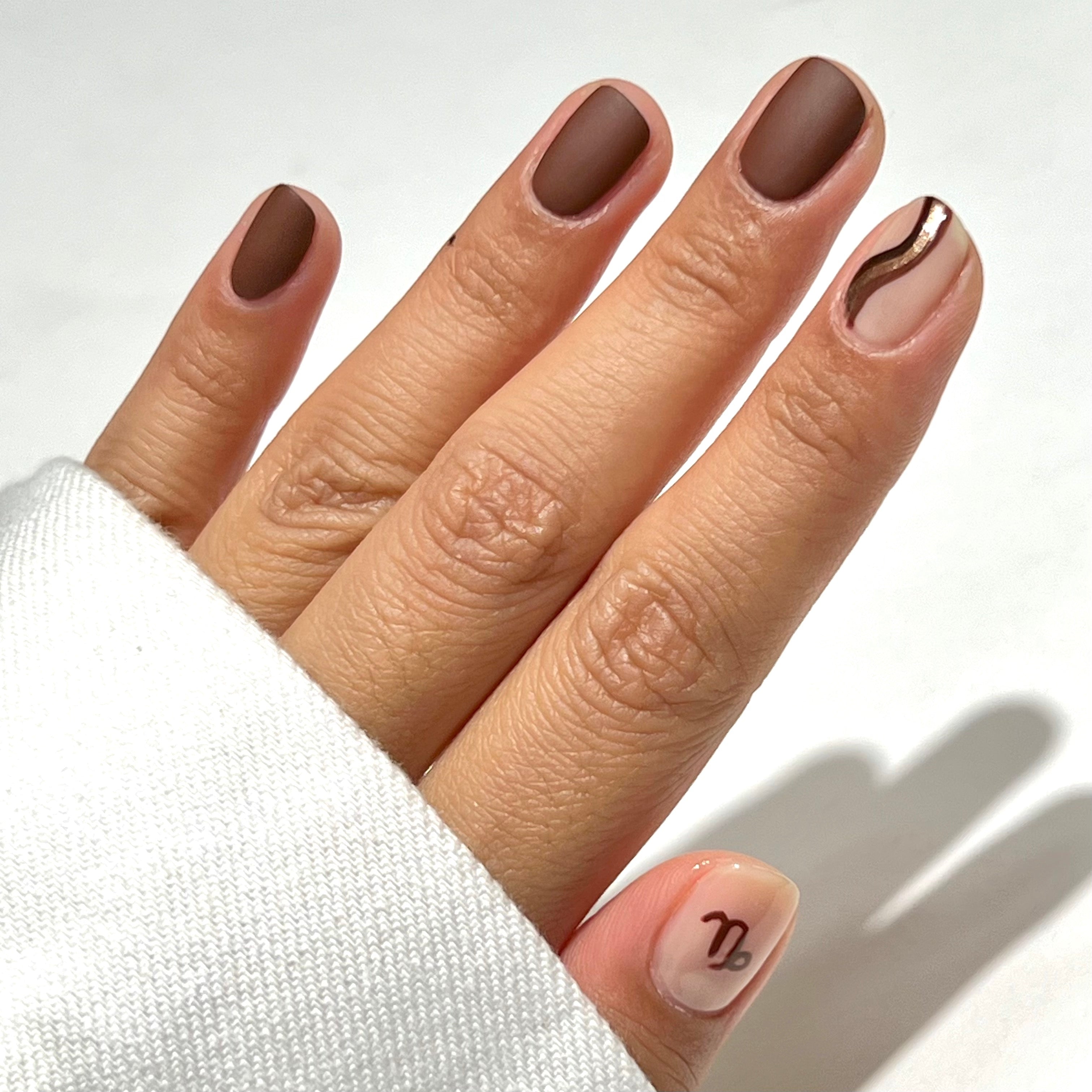 A hand with short, manicured nails. Most nails are painted matte brown; the ring finger has a nude base with a brown wavy line, and the thumb is nude with a brown Capricorn zodiac symbol. The person wears a white sleeve.