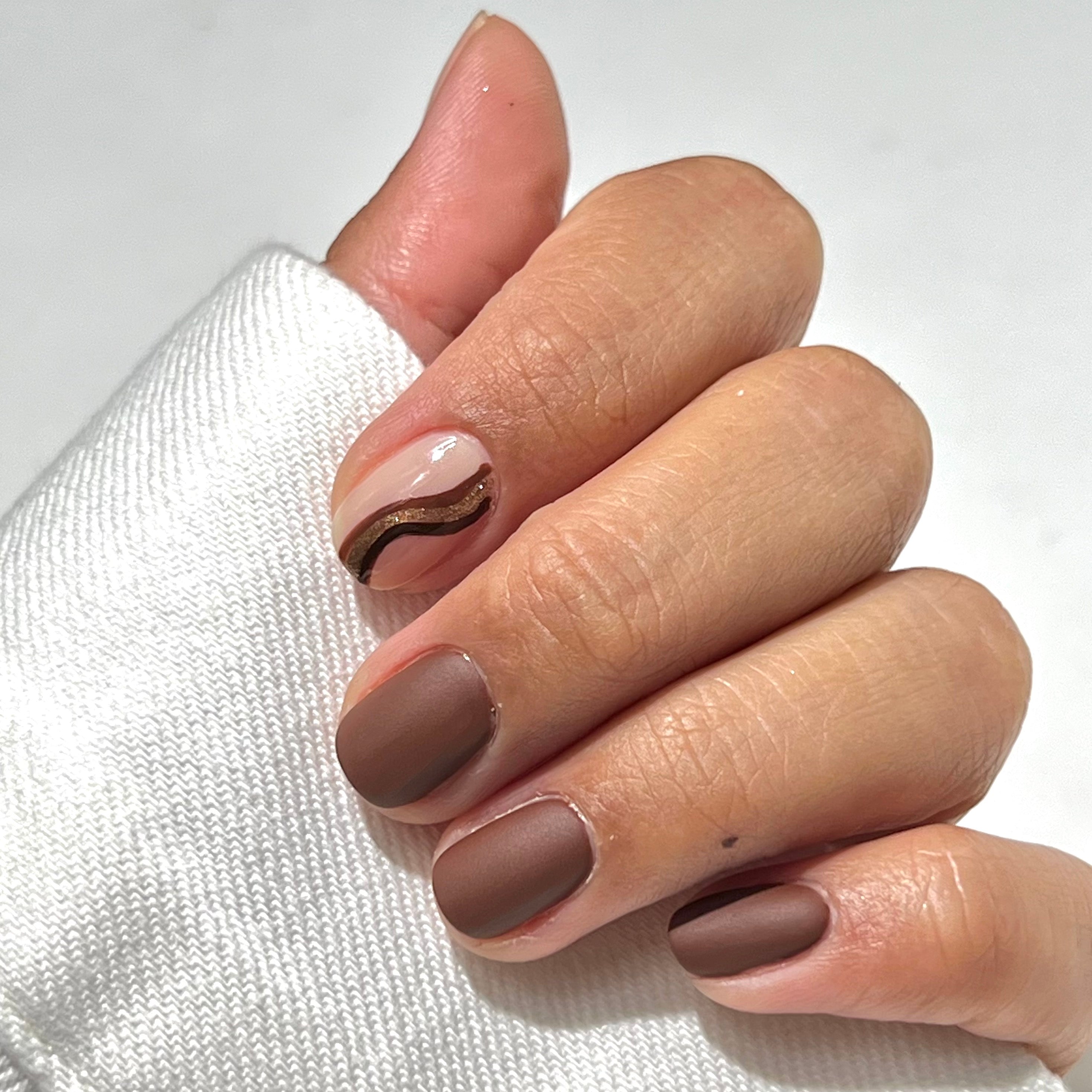 A hand with short, neatly manicured nails. Four nails are painted matte brown, while the index nail is nude with wavy brown and gold lines. The hand is partially covered by a white fabric sleeve.