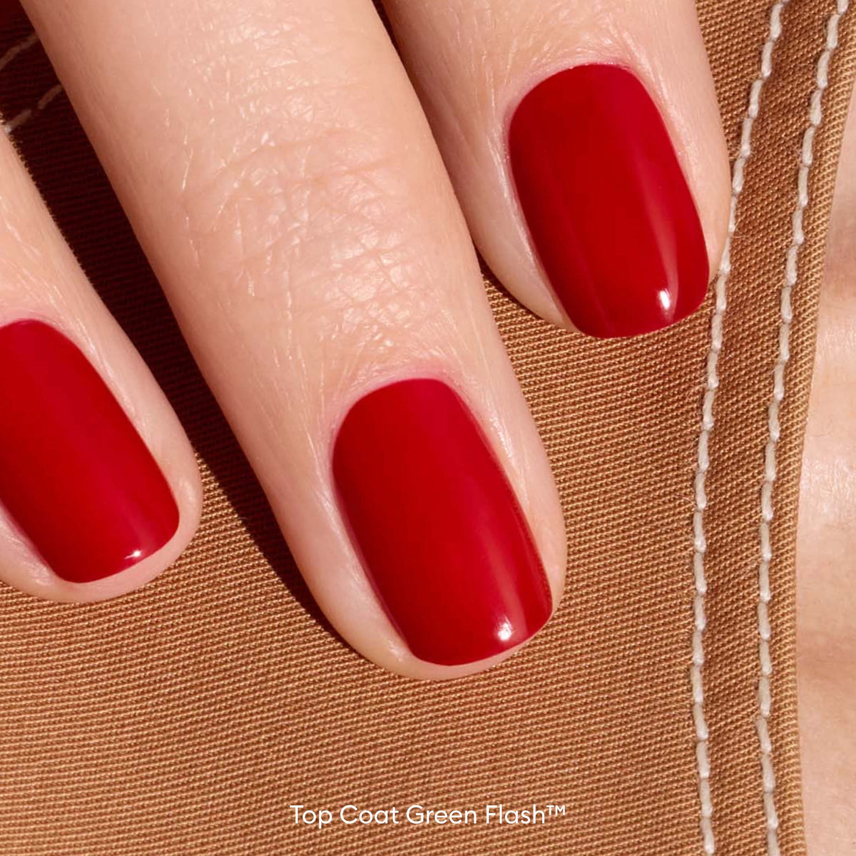 A close-up of three fingers with neatly manicured, glossy red nails resting on tan fabric with visible white stitching. The words Top Coat Green Flash™ appear at the bottom of the image.