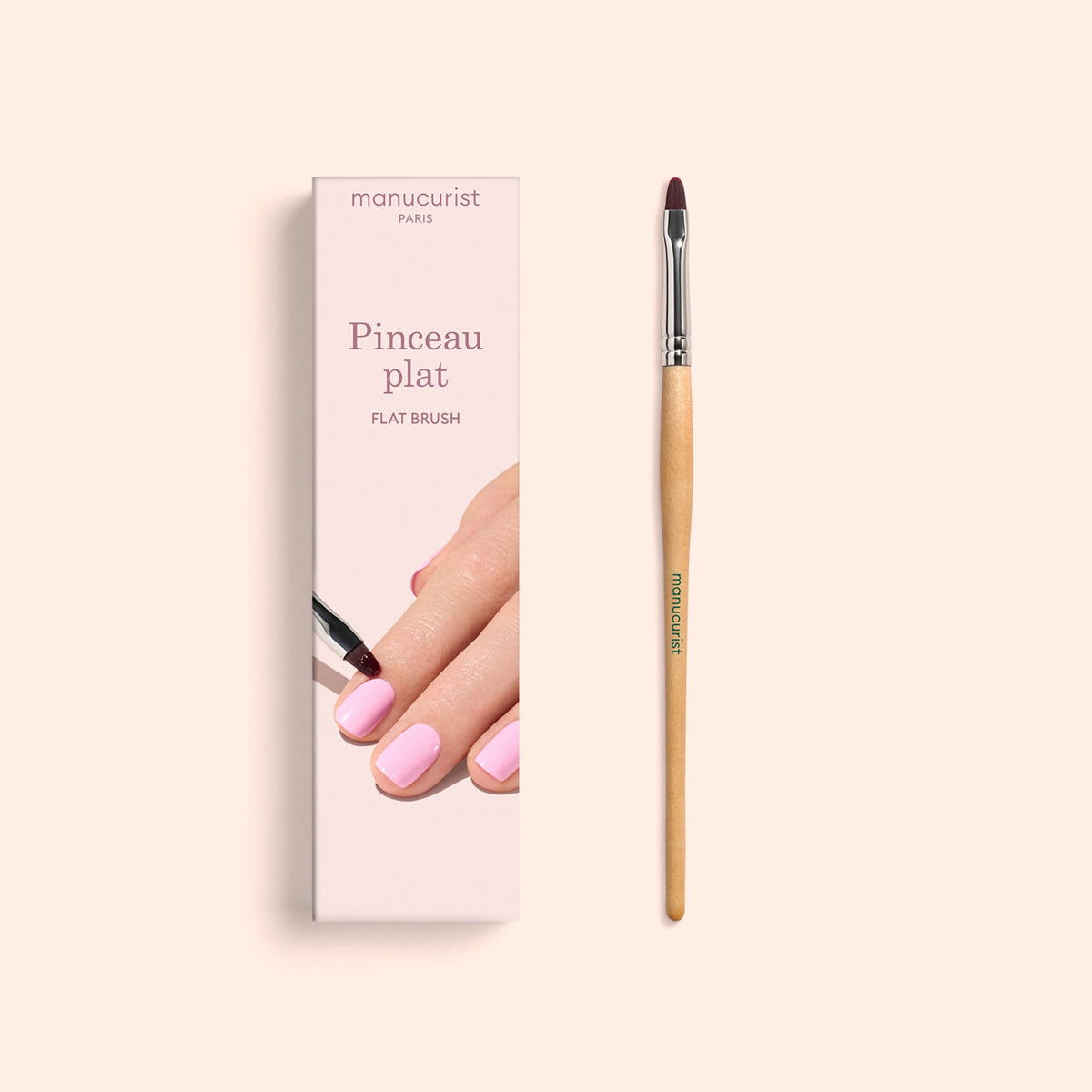 Un pinceau Flat Brush Manucurist au manche en bois, idéal pour une manucure précise et le soin des cuticules, posé à côté de sa boîte ornée d&