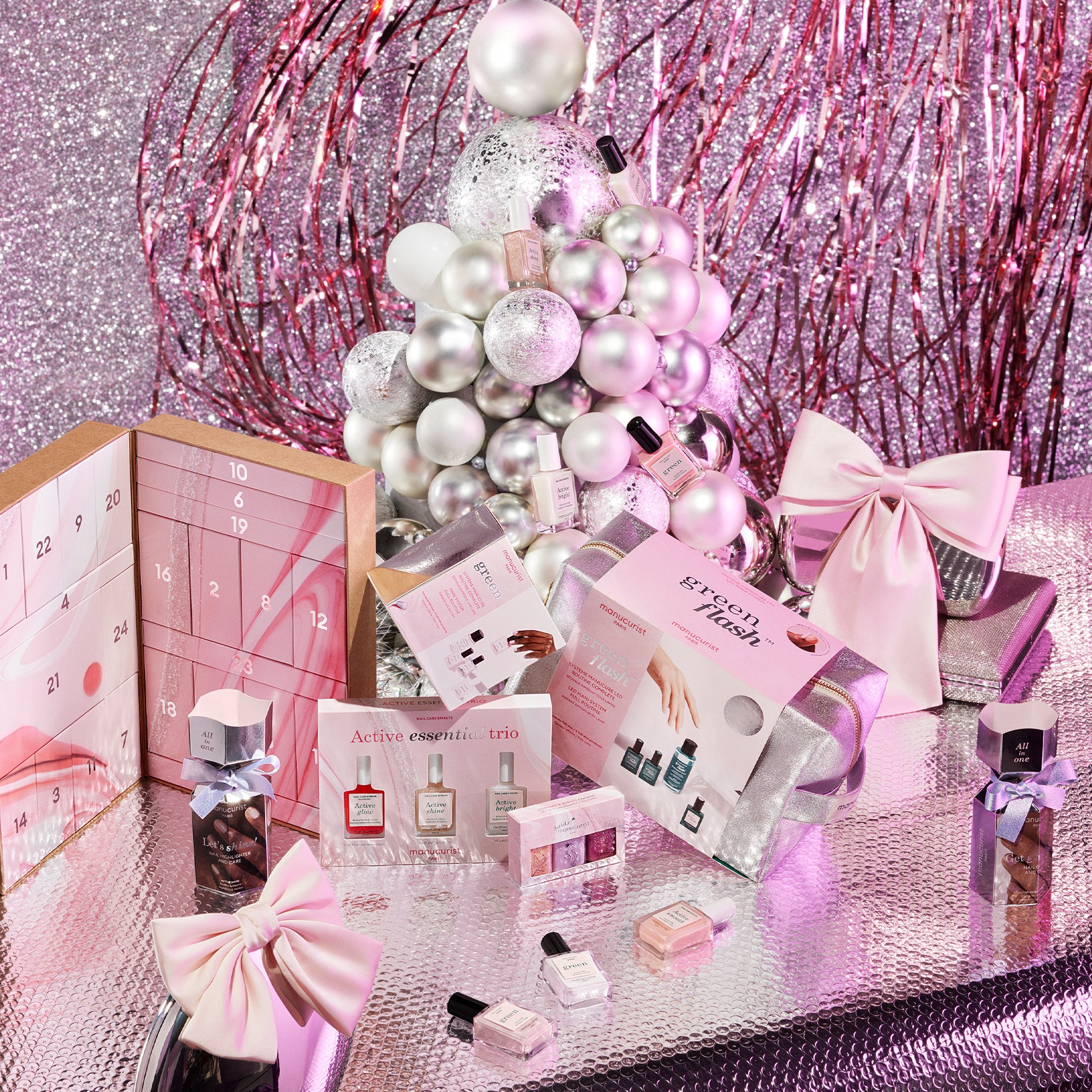 A festive display featuring a white and silver ornament Christmas tree, beauty gift sets, perfume bottles, and pink ribbon-wrapped presents, set against a sparkly pink and silver backdrop.