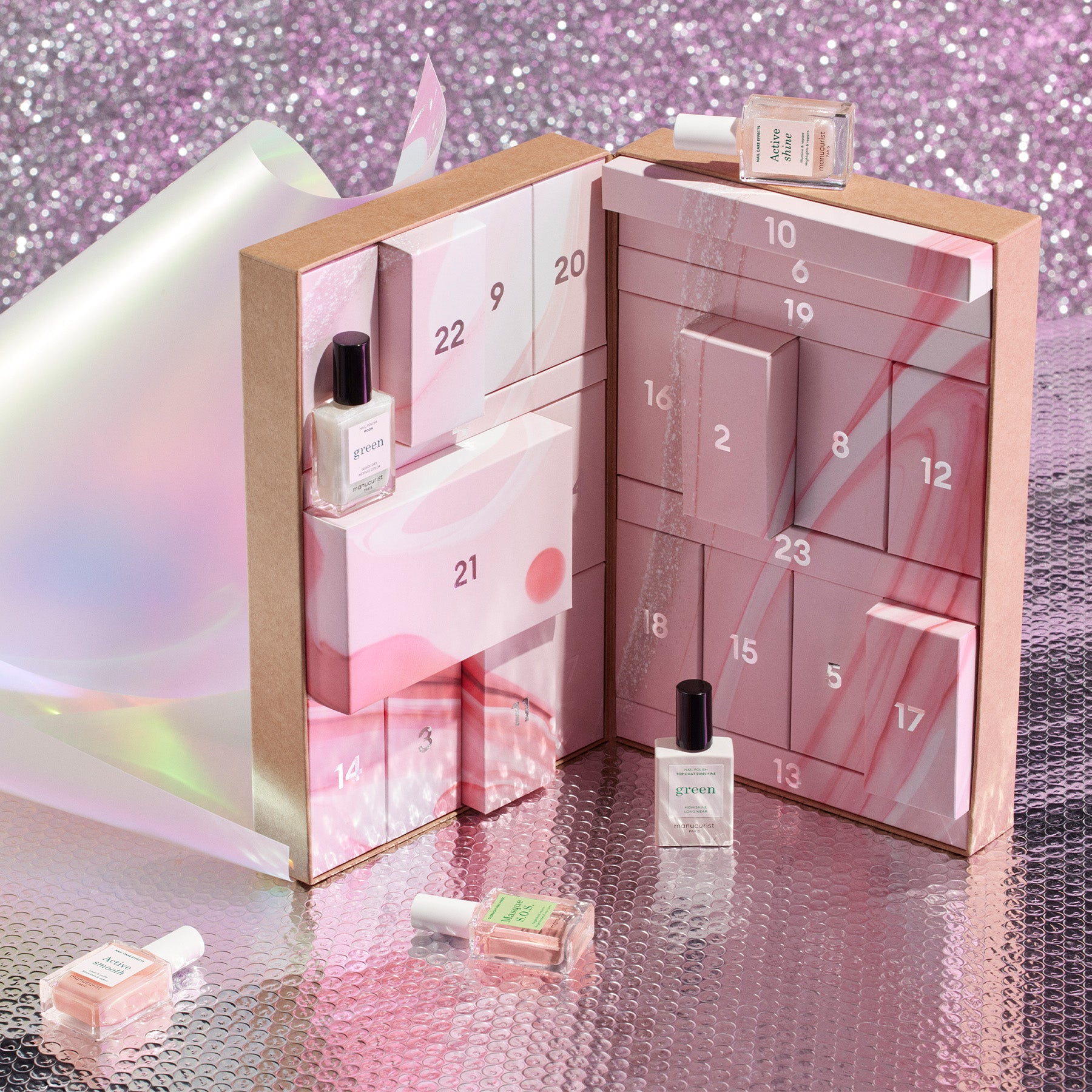 An open advent calendar with pink and marble-patterned numbered drawers, each containing small nail polish bottles. Three bottles are displayed outside the calendar. The background is iridescent and sparkly.