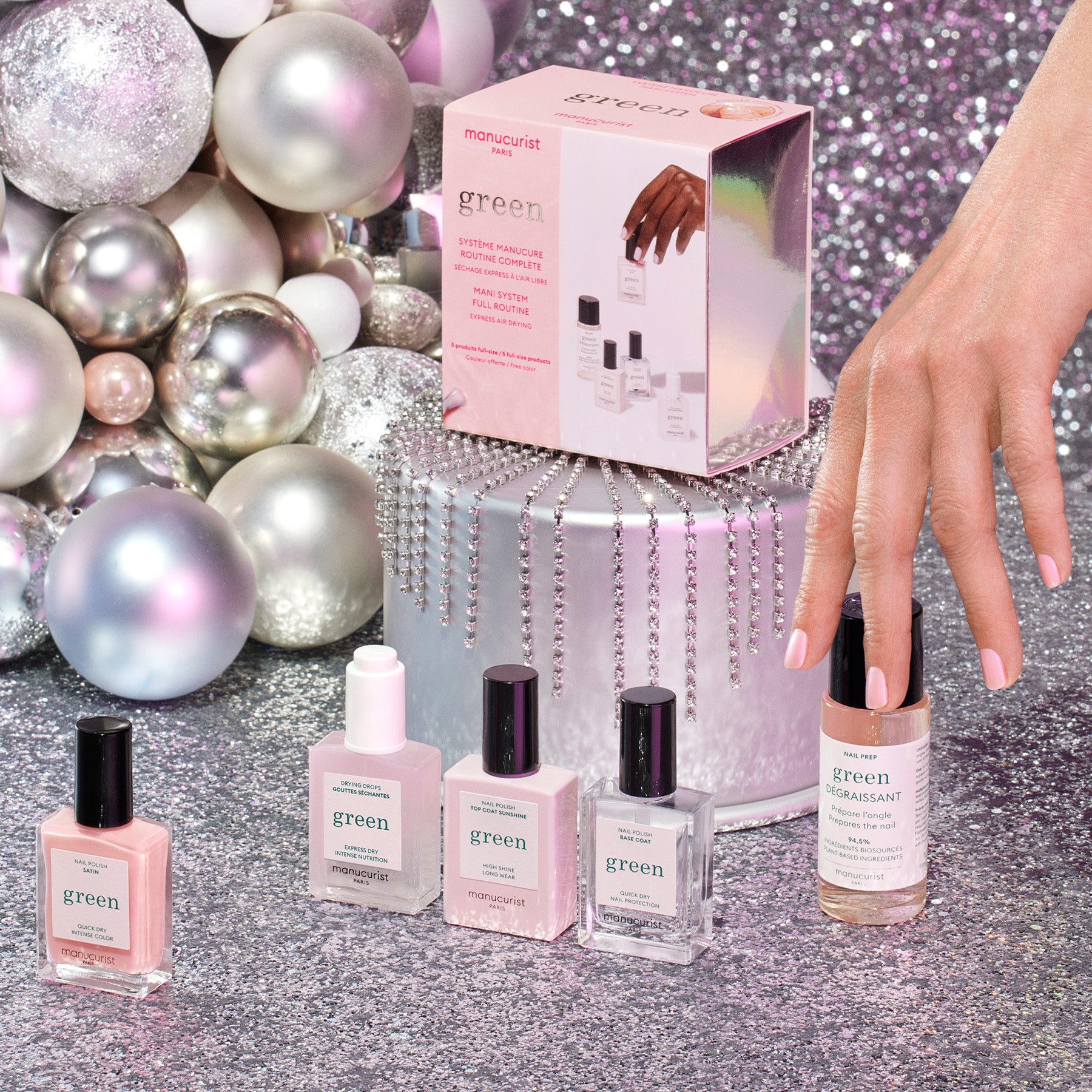A hand with pink nail polish reaches for a nail polish bottle in front of a silver gift box. Five nail polish bottles and a product box are displayed among shiny silver, white, and pink holiday ornaments.