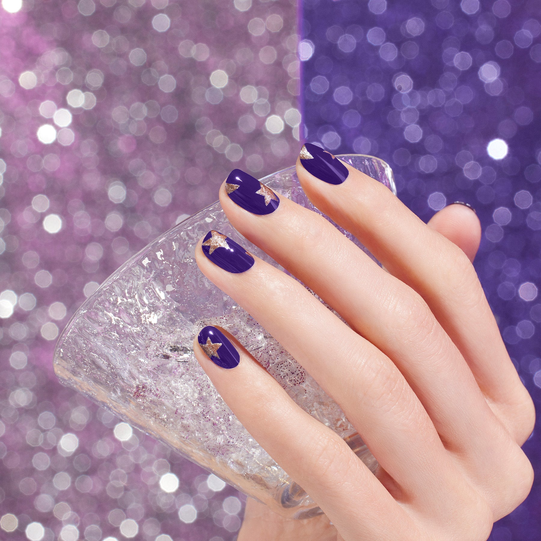 A hand with purple nail polish and gold star designs holds a textured, clear glass against a sparkly purple background.