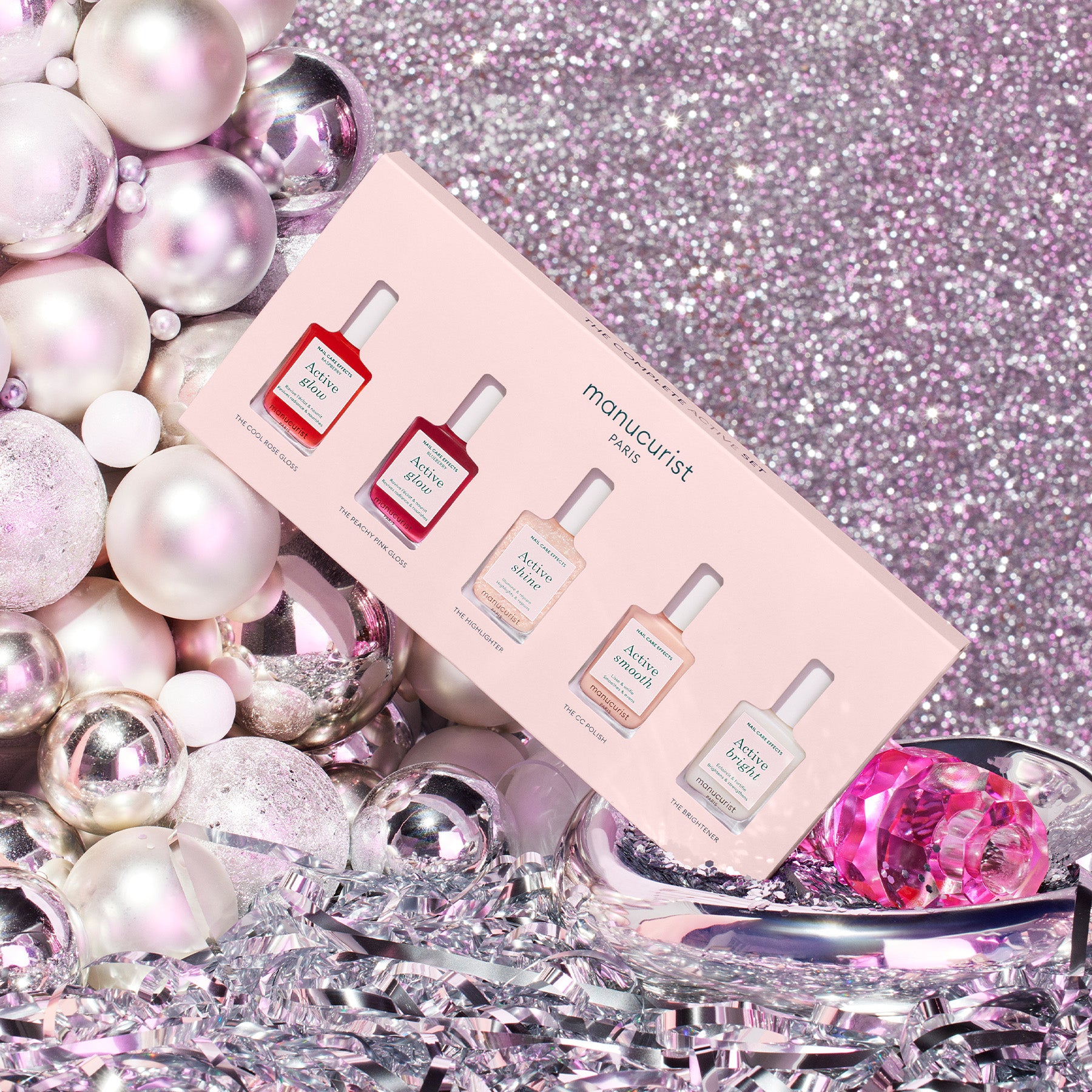 A set of five Manucurist Paris nail polishes in a pink box is displayed among shiny silver and pink ornaments and tinsel on a sparkling, glittery pink background.