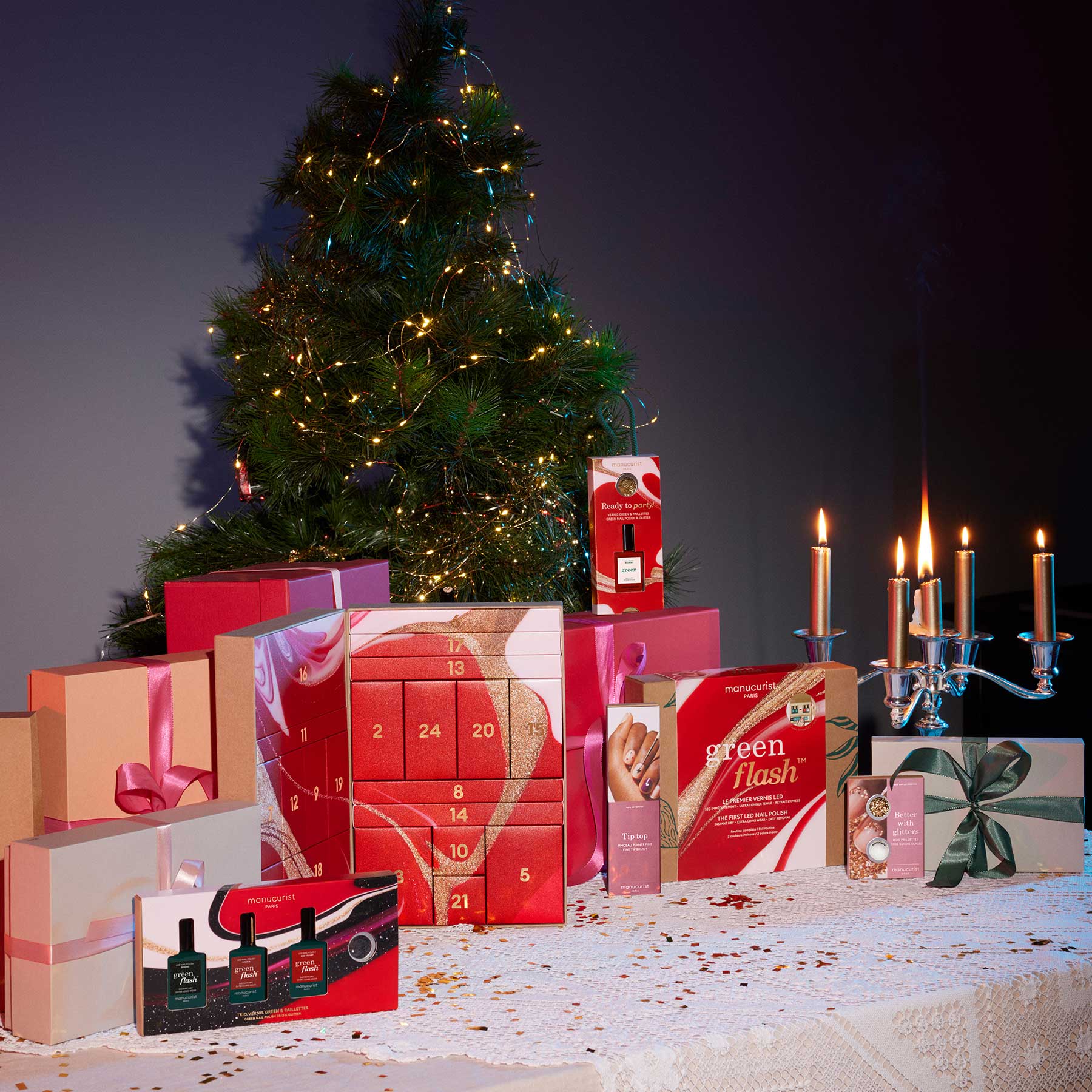A decorated Christmas tree stands behind a table covered with wrapped gifts, a red advent calendar, beauty product boxes, and a lit silver candelabra, creating a festive holiday scene.