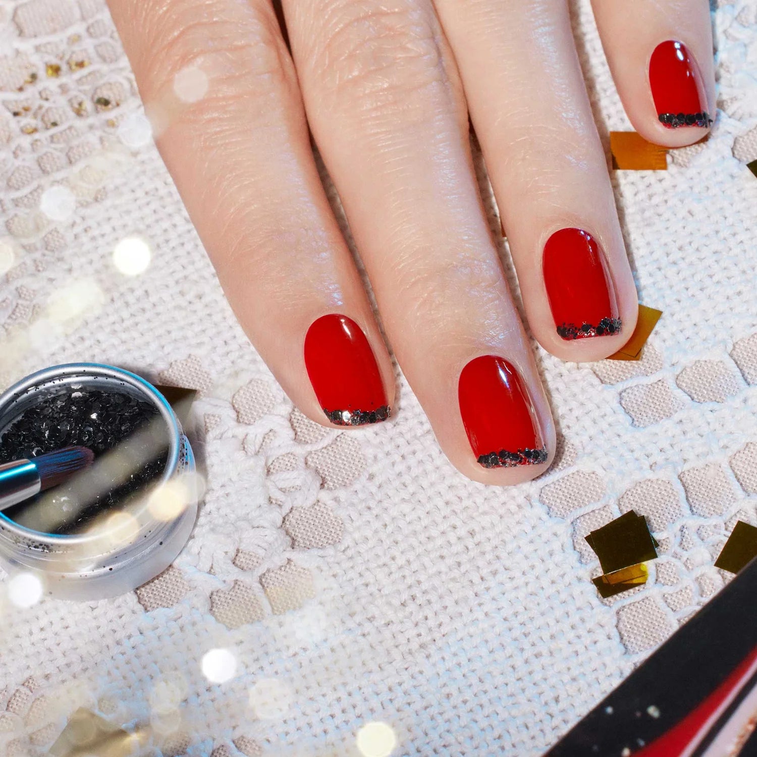 A hand with red nail polish and black glitter tips rests on a white lace fabric. A small container of black glitter and a brush are nearby, along with some gold confetti pieces.