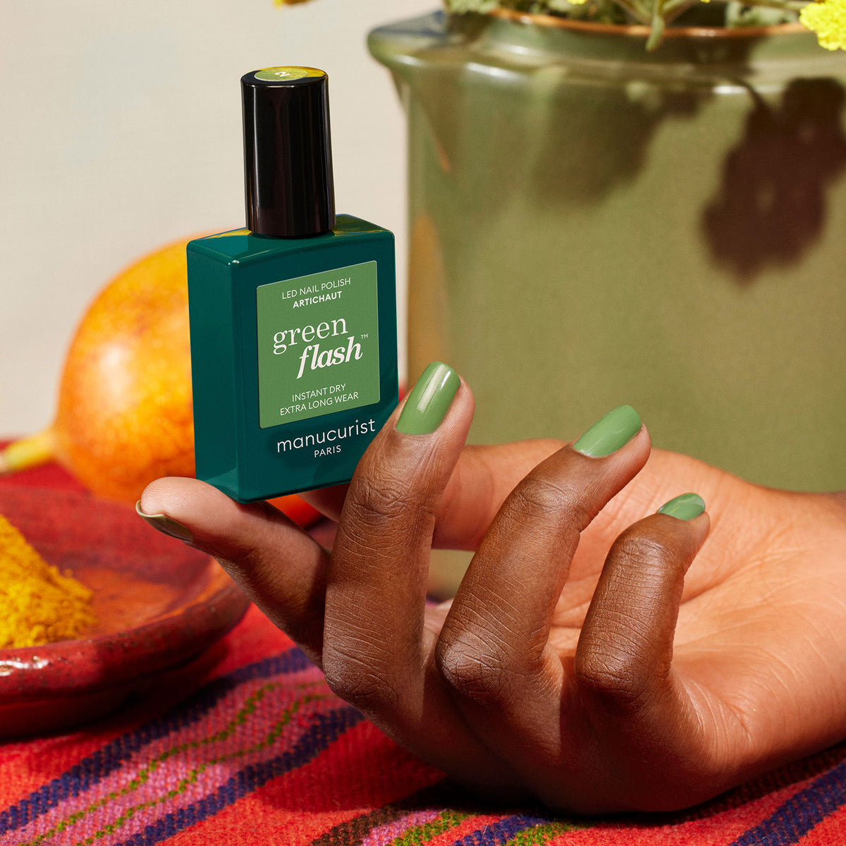 Une main aux ongles peints en vert tient un flacon de vernis Manucurist Artichoke. Le décor évoque un jardin marocain, avec un tissu aux couleurs vives, un bol de fruits et un vase vert en arrière-plan.
