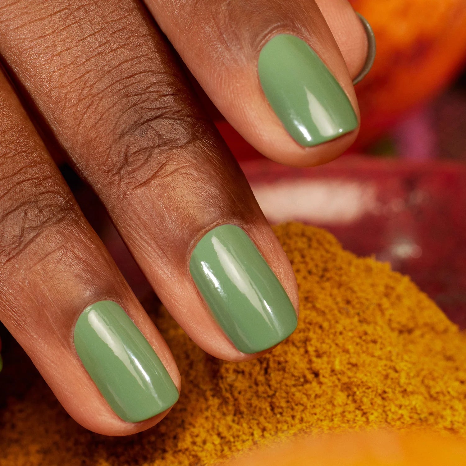 A close-up of a hand with dark skin and neatly manicured nails painted with glossy, light green nail polish. The background includes a soft blur of yellow and red colors.
