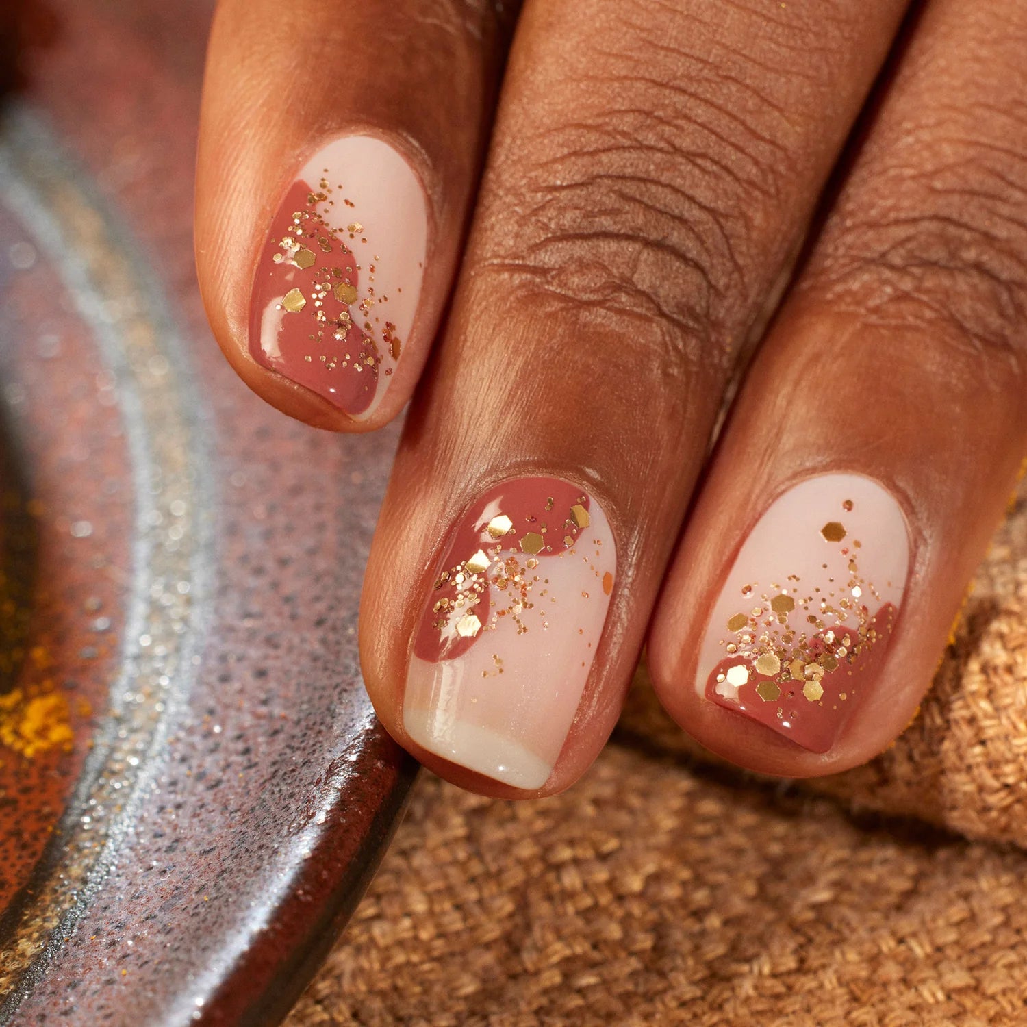 Close-up of a hand with nails painted in a nude and pink gradient, topped with gold glitter. The fingers rest near a textured brown surface and a ceramic dish.