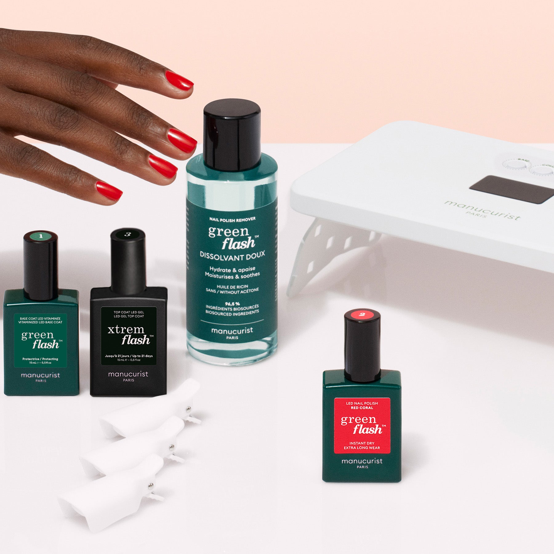 A hand with red-painted nails reaches toward the Manucurist Discovery Kit, which includes Green Flash gel polish, vitamin-rich base coat, remover, nail clips, and a UV lamp, all arranged on a white surface.