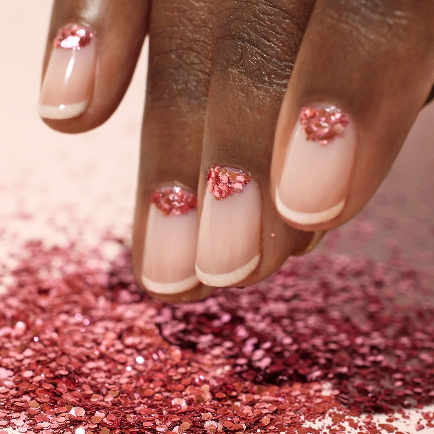 A hand with natural, nude-colored nails features French tips and pink glitter clusters near the cuticles. Pink glitter is scattered on the surface beneath the fingers.
