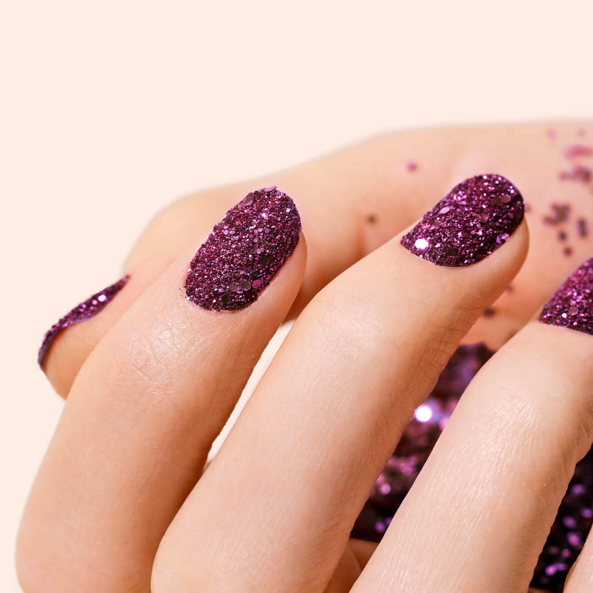 Gros plan d’une main aux ongles vernis d’un gel pailleté violet. Des paillettes biodégradables Bordeaux de Manucurist sont dispersées sur la peau et le fond beige doux.