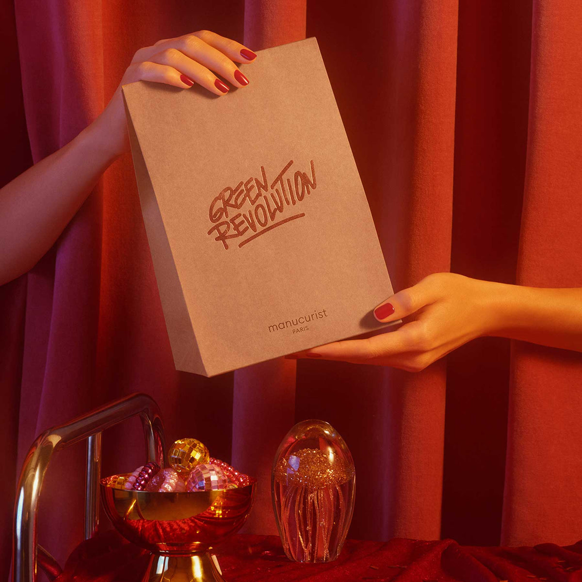 Deux mains aux ongles vernis de rouge tiennent un Small Gift Bag beige étiqueté Manucurist devant des rideaux rouges. Le cadeau de soin des ongles brille, et en dessous, une décoration en verre et une coupe de boules scintillent sur la surface en velours rouge.