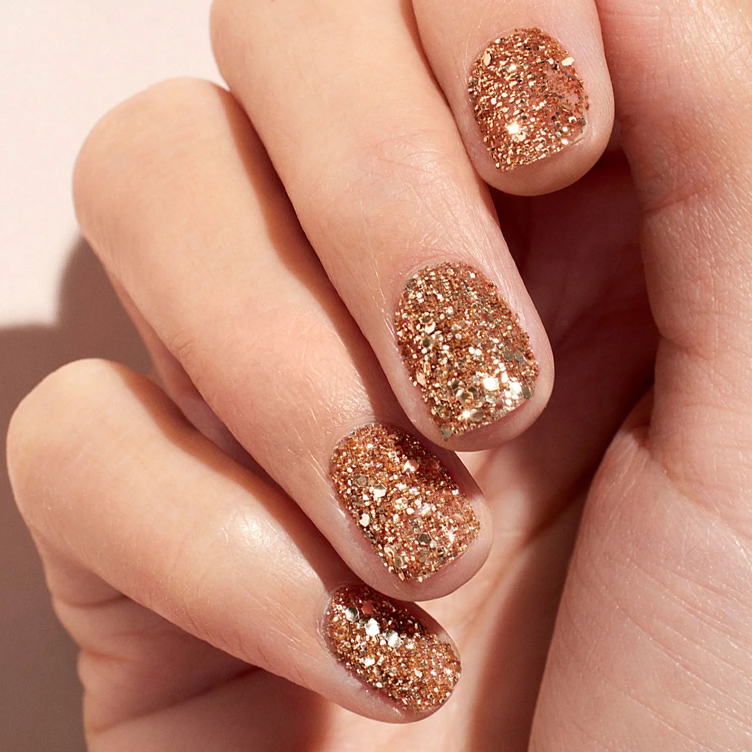 A close-up of a hand with neatly manicured nails painted in a sparkly gold glitter polish. The nails are short and rounded, and the glitter reflects light, creating a shimmering effect.