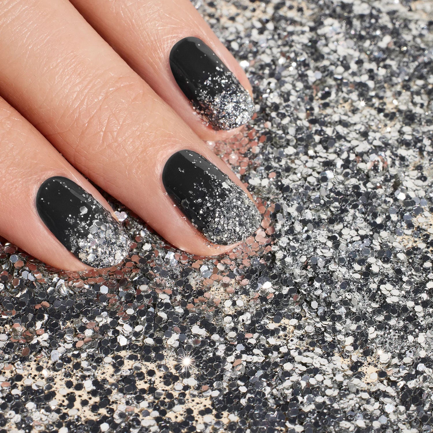 A close-up of a hand with black nail polish and silver glitter ombré, resting on a surface covered in shiny silver glitter.