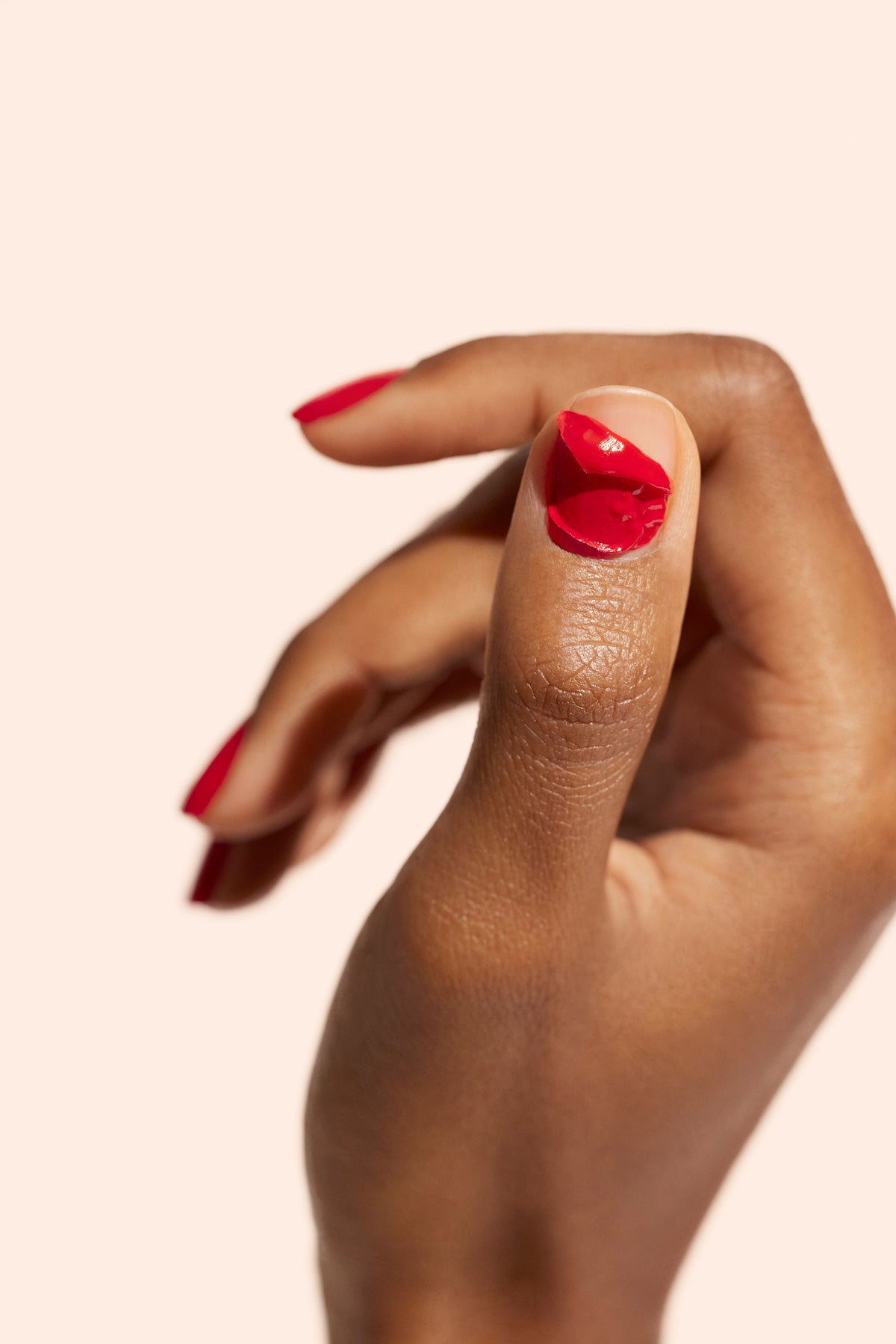 Une main à la peau brun moyen montre un pouce avec du vernis à ongles rouge écaillé. Les autres ongles sont également peints en rouge. L’arrière-plan est d’une couleur beige clair unie.