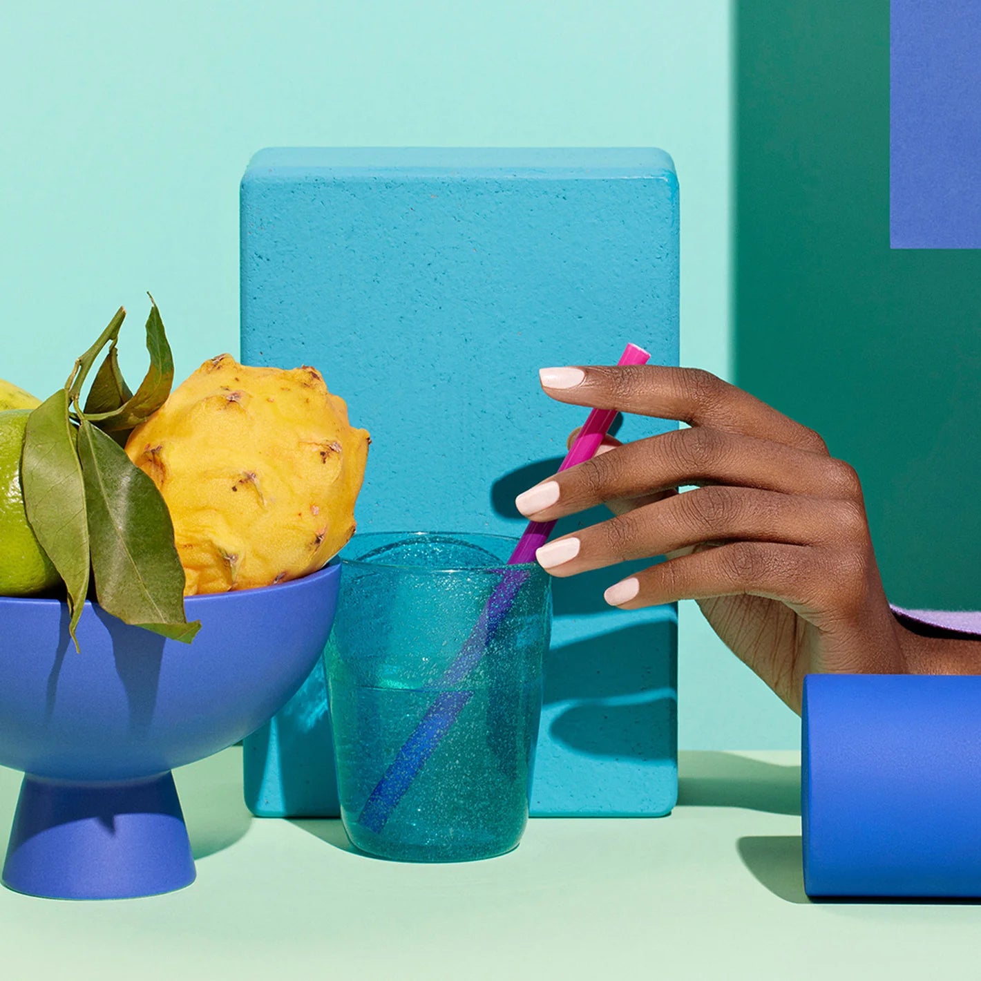 A hand with pale pink nails holds a pink straw in a blue glass. Next to it is a blue bowl of fruit, including a yellow pitaya and green pears. The background features teal and blue blocks on a turquoise surface.