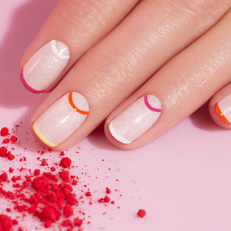 A close-up of a hand with light pink, shimmery nail polish featuring colorful, curved lines at the base of each nail. Red powder is sprinkled on the pink surface nearby.