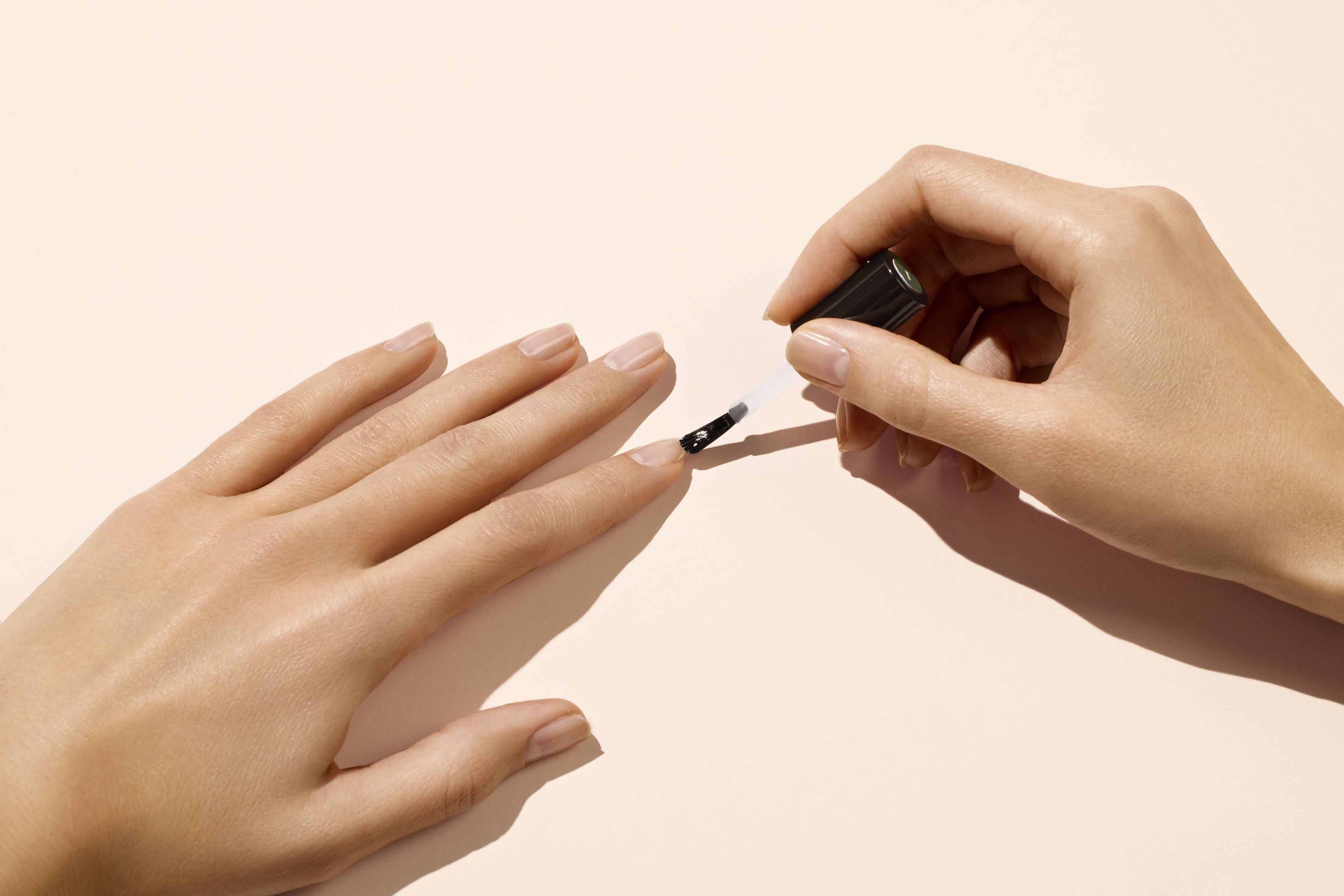 A person applies clear nail polish to the fingernails of their left hand using a brush, with both hands resting on a light beige surface.