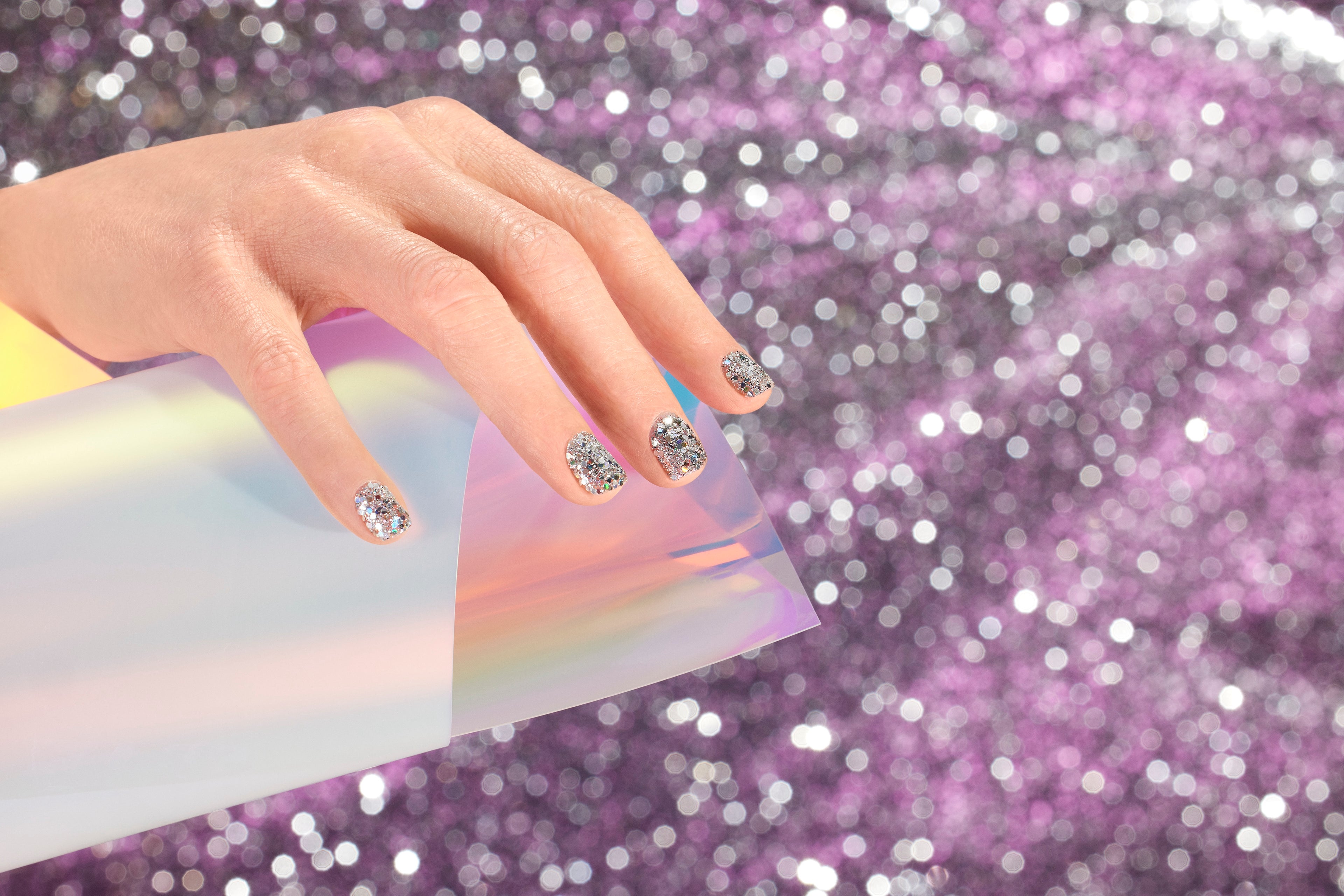A hand with silver glittery nail polish holds a piece of iridescent paper against a sparkling purple and silver bokeh background.