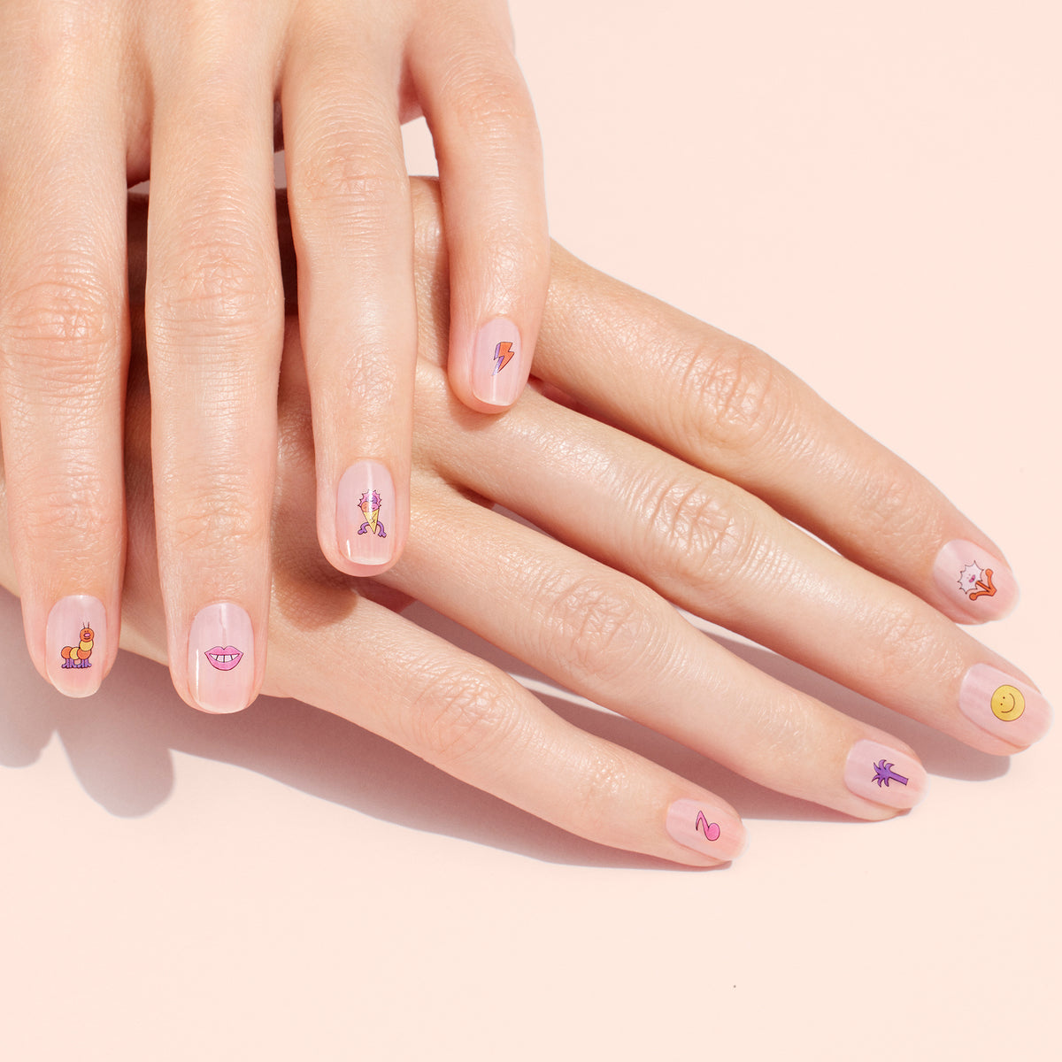 Close-up of hands with short, natural nails, each decorated with a different playful Summer Nail Art Sticker from Manucurist—smiley faces, lips, and more—over a light pink base for a fun manicure.