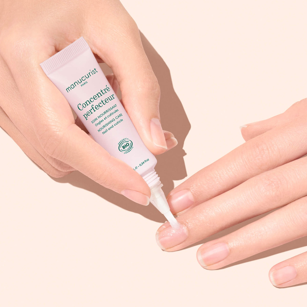 A person uses the Manucurist Nourishing Nail Pen - Cosmos Organic on their fingernails, showcasing nail and cuticle care with clean, manicured hands against a light peach background.