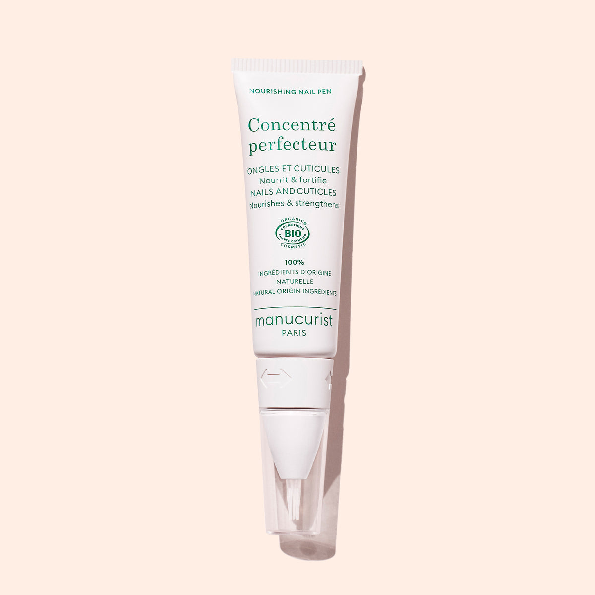 Un tube blanc de Manucurist Nourishing Nail Pen - Cosmos Organic avec du texte vert se tient debout sur un fond rose clair — idéal pour le soin des ongles et des cuticules.