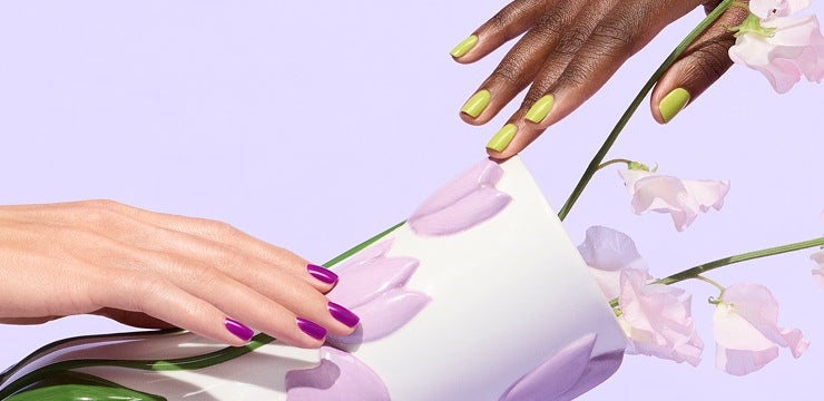 Two hands with manicured nails—one with lime green polish, the other with bright purple—touch a white vase decorated with purple tulip designs, holding light pink flowers against a pastel lilac background.