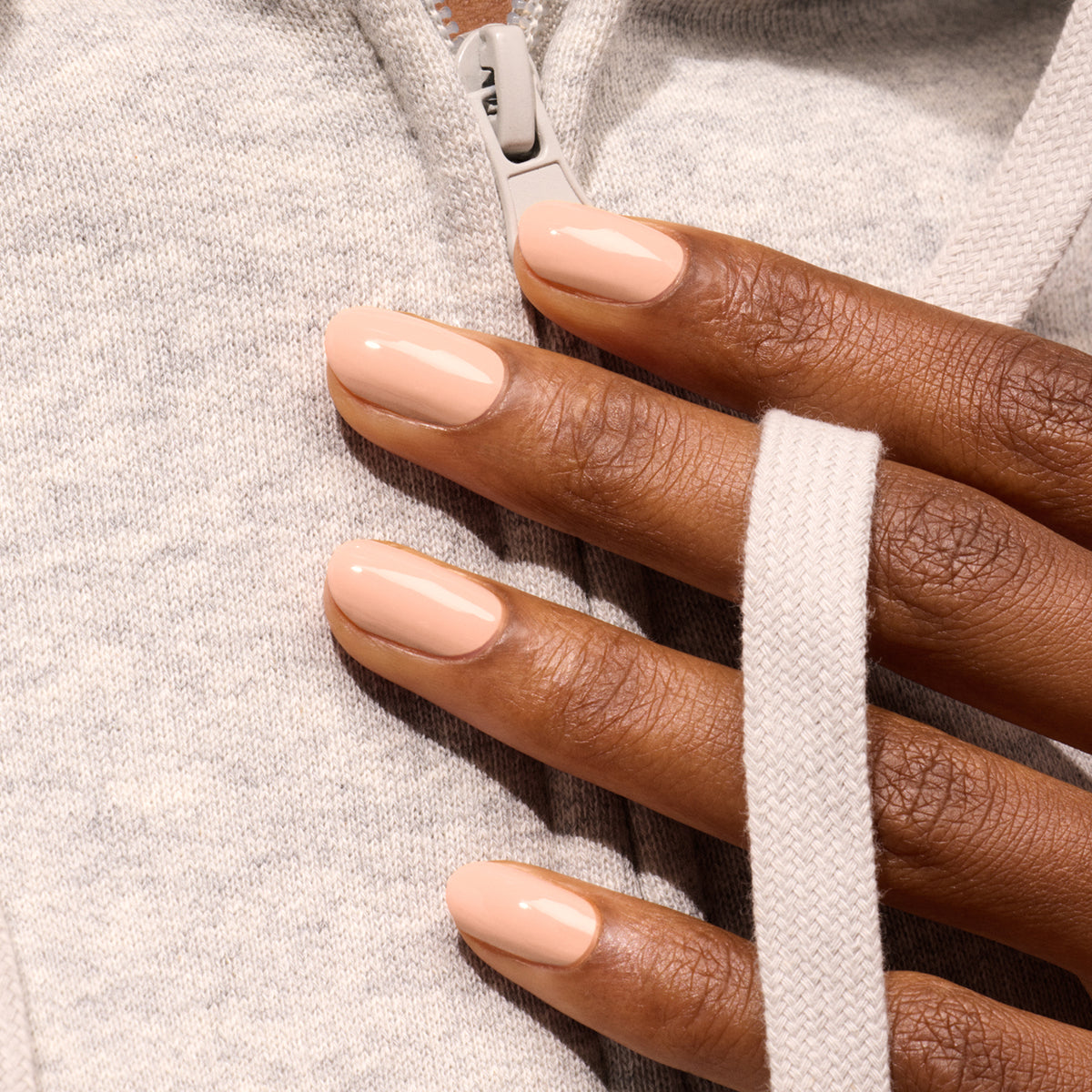 A hand with neatly manicured nails painted in Pink Sand by Manucurist rests on a light gray zip-up hoodie, with fingers holding one of the hoodie’s white drawstrings.