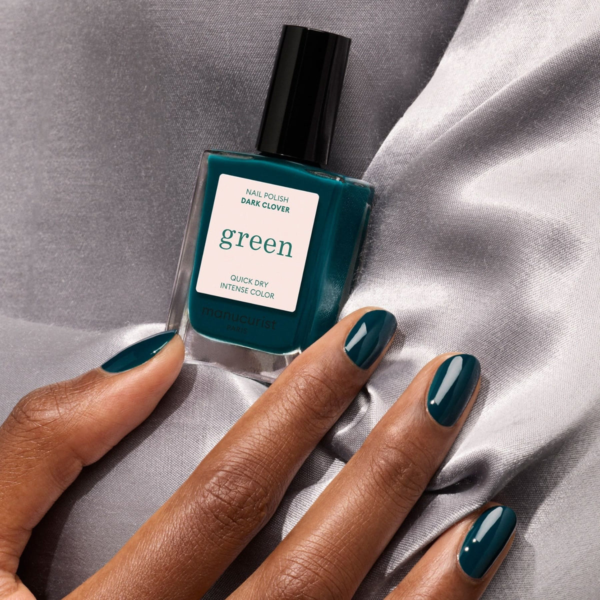 A hand with dark teal nails holds a Manucurist Dark Clover nail polish bottle, resting on gray satin fabric.