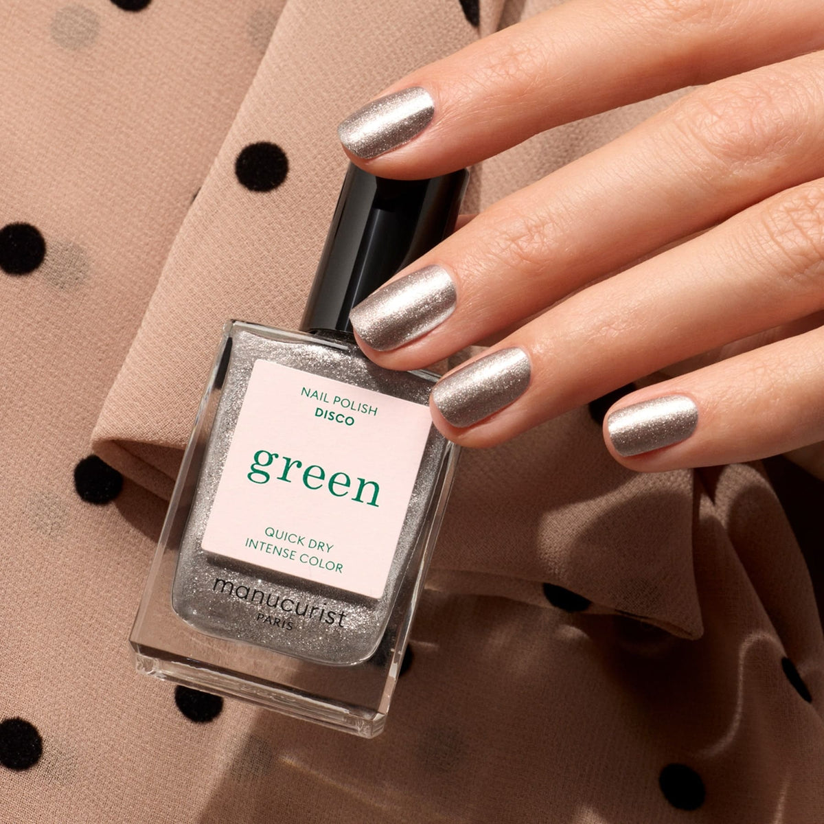 Une main aux ongles argentés opaques et pailletés tient un flacon de Disco de Manucurist, sur un tissu beige à pois noirs.