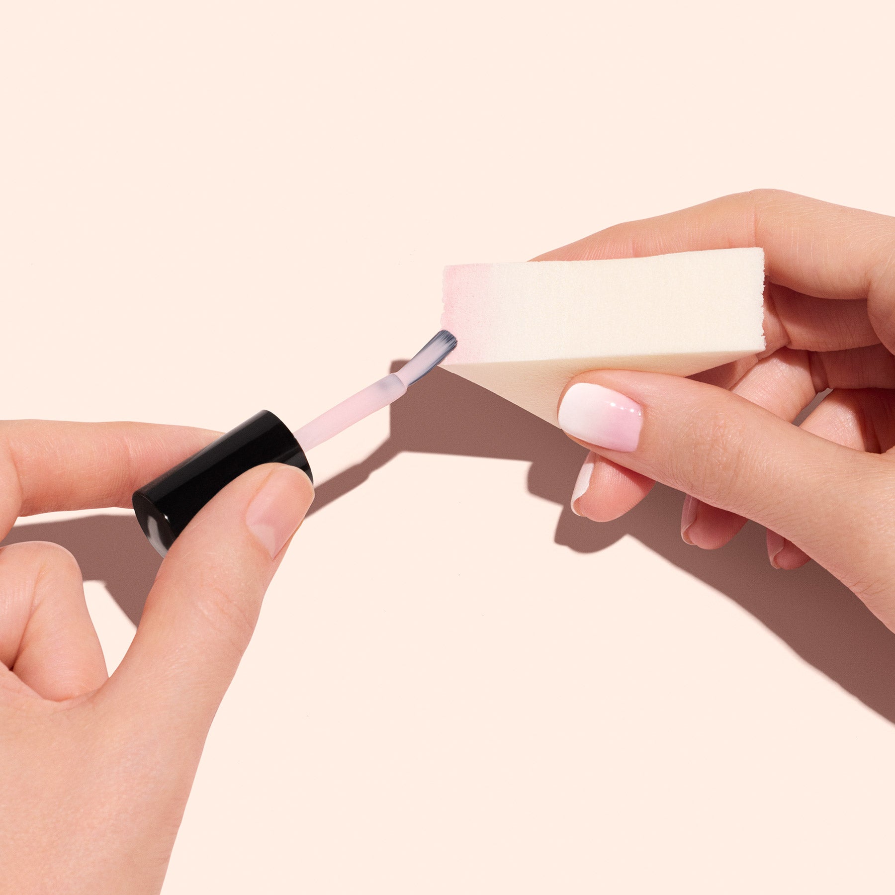 A person holds a Manucurist Nail Art Sponge (x4) in one hand and applies light pink polish to it with a brush. Their neatly manicured Baby Boomer nails are shown against a light beige background.