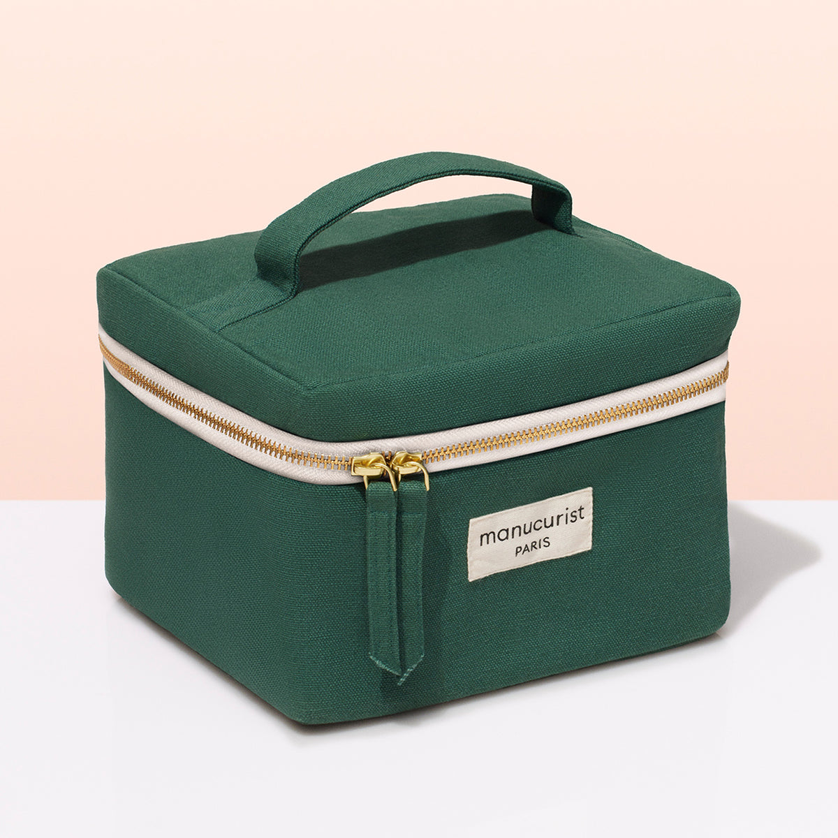 Un vanity case Manucurist-test en tissu vert avec une anse, des fermetures éclair dorées et une étiquette beige "manucurist PARIS" — idéal pour ranger ses vernis ou son matériel de manucure — présenté sur une surface blanche sur fond rose clair.
