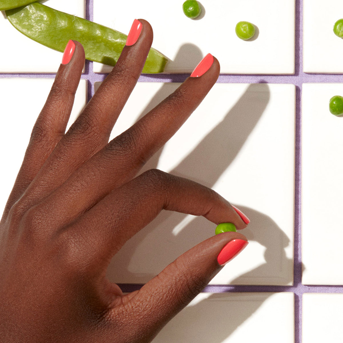 Une main portant le vernis gel rose corail Pulp de Manucurist tient un petit pois au-dessus de carreaux blancs, entourée d&