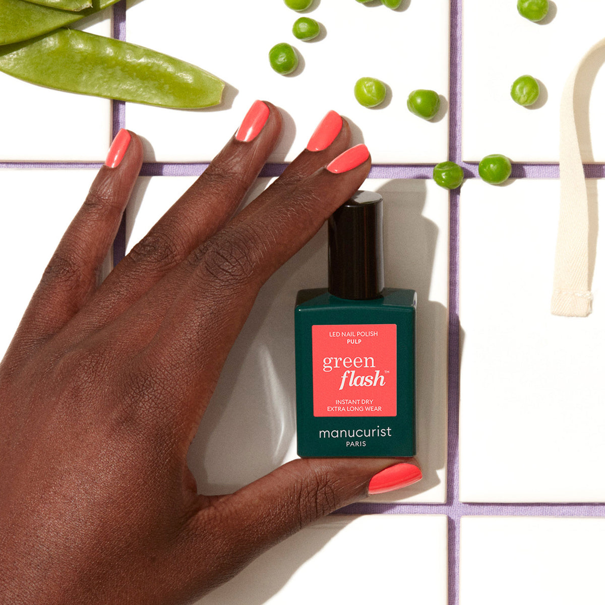 Une main avec un vernis gel rose corail tient un flacon Manucurist Pulp sur une surface carrelée blanche, entourée de petits pois verts et de cosses de pois gourmands.