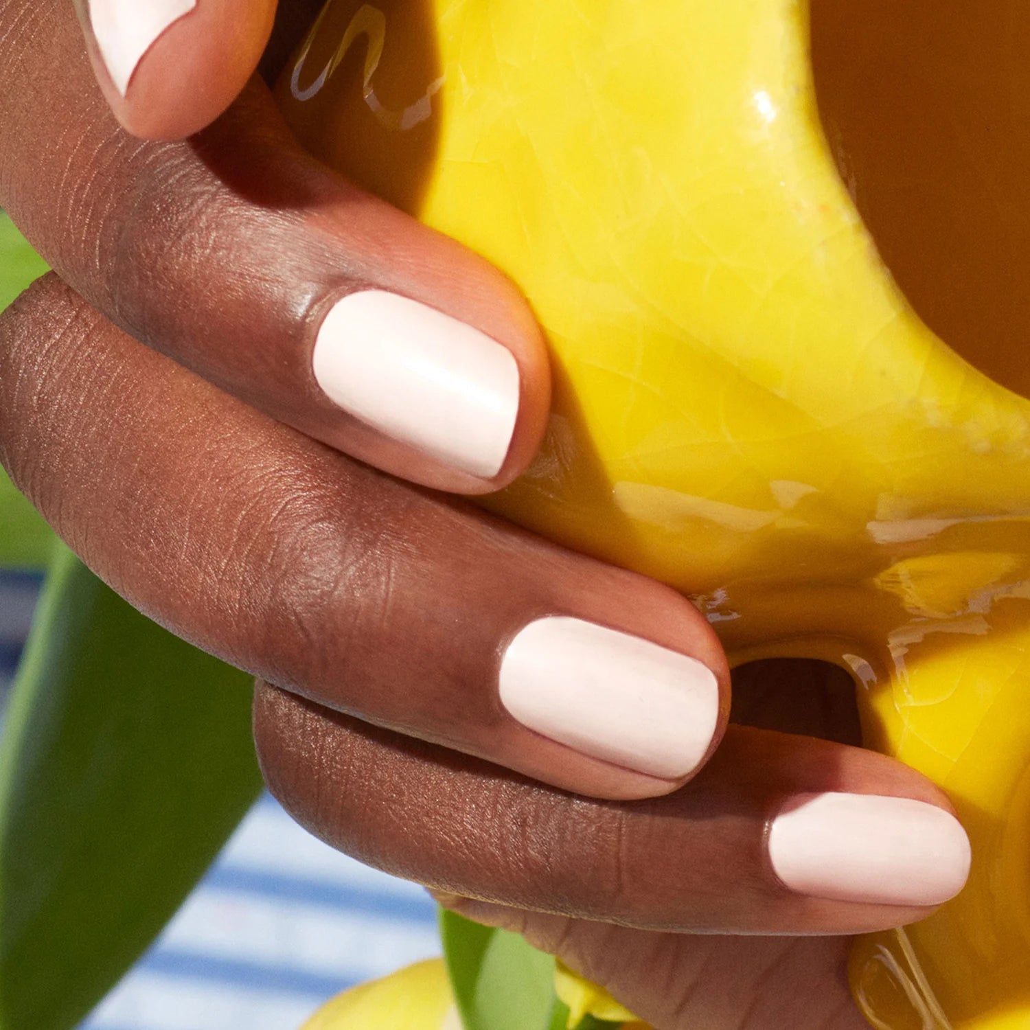 A close-up of hands with light pink nail polish holding a bright yellow ceramic object, with green leaves and a blue-and-white striped background visible.