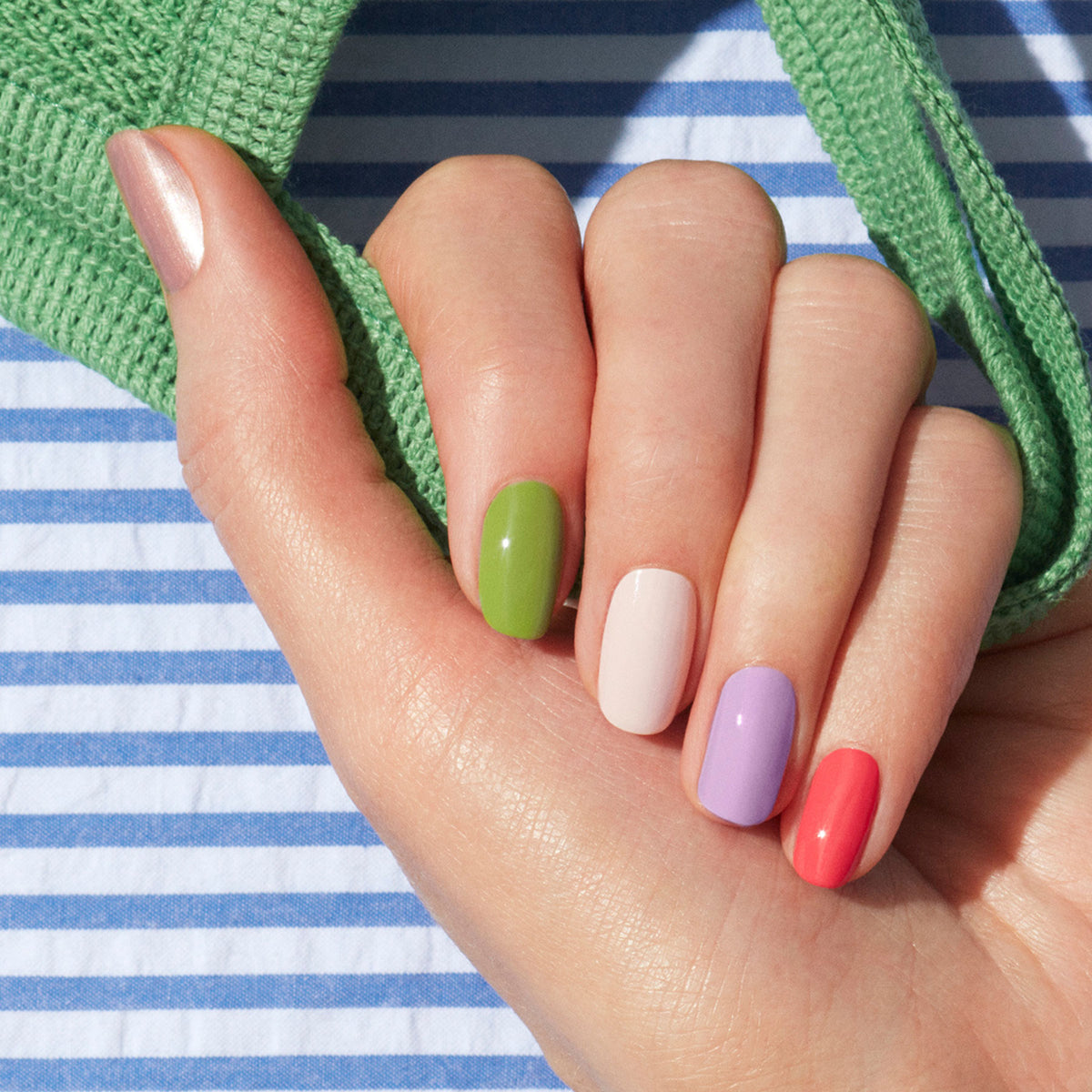 Une main aux ongles impeccablement manucurés, vernis de cinq couleurs — rose métallisé, vert, blanc, lavande et corail —, repose sur un tissu rayé avec une lanière verte, mettant en valeur un nail art ludique réalisé avec le Dot it better! de Manucurist.