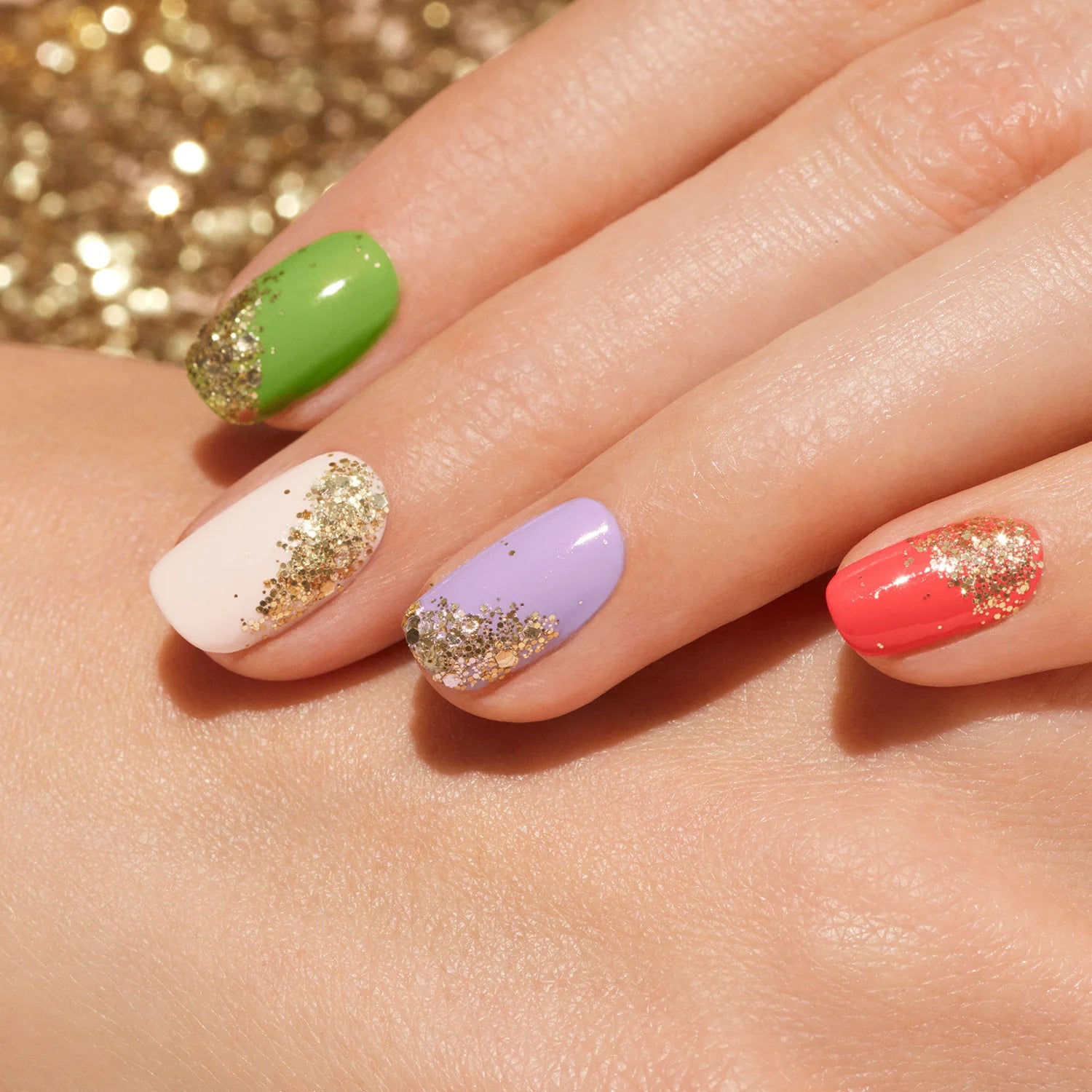 A hand with nails painted green, white, lavender, and coral, each accented with gold glitter at the base, resting on another hand against a sparkling gold background.
