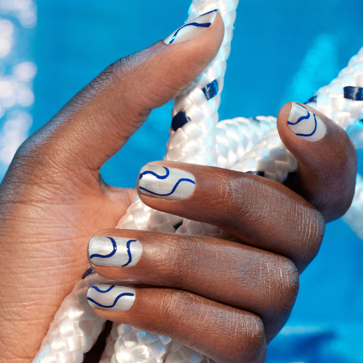 Une main aux ongles bleu clair avec un nail art ondulé bleu foncé — vernis avec Midnight Swim de Manucurist issu de la collection Life Aquatic — tient une corde blanche avec des détails bleus sur un fond bleu vif.