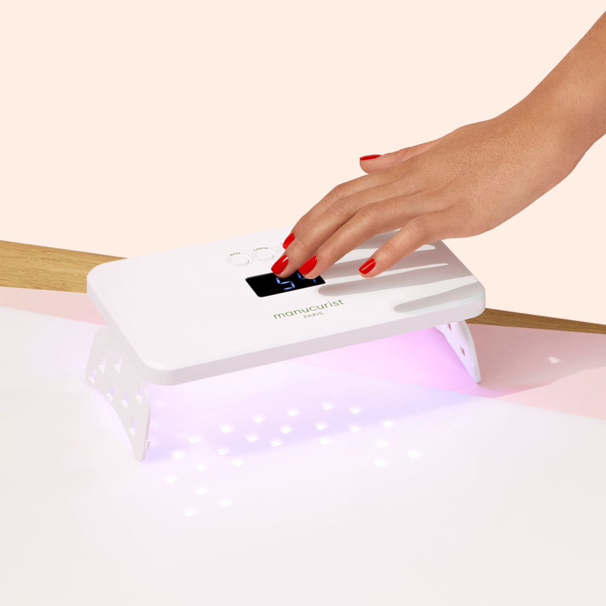 A hand with red Green Flash gel polish cures under the Manucurist Green Flash™ Premium 36W Lamp, which emits purple light for quick drying. The scene features a minimal, light background.