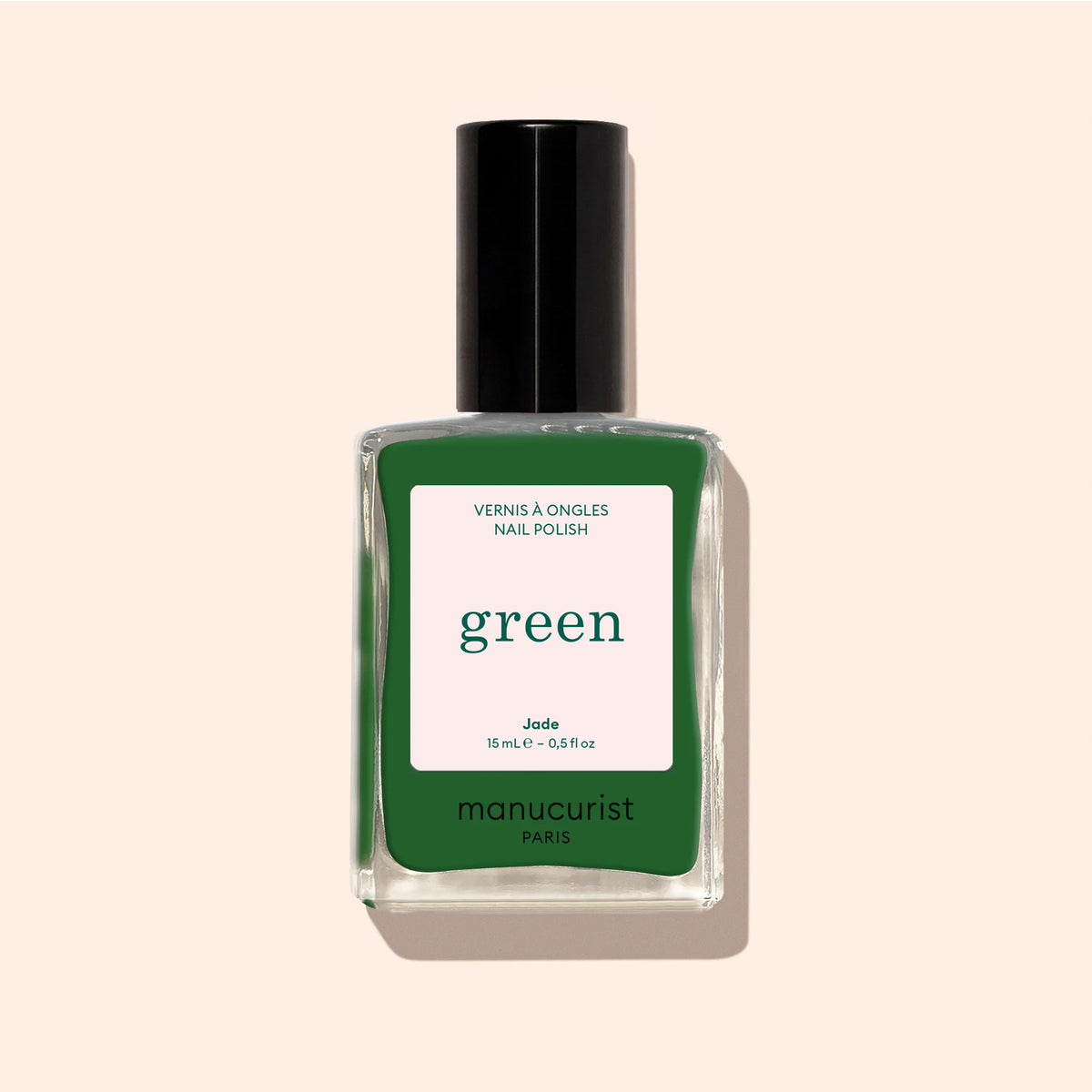 Un flacon de vernis à ongles Manucurist Jade vert minéral aux sous-tons froids, dans une teinte vert foncé, avec un bouchon noir et une étiquette beige, est présenté sur un fond beige clair.
