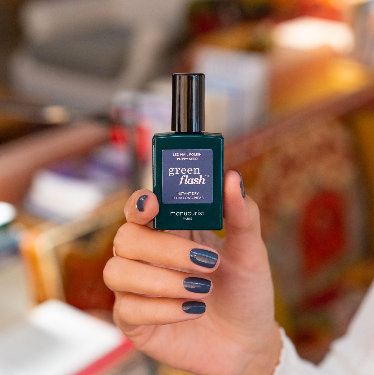 Une main aux ongles soigneusement manucurés, peints dans la chic teinte Poppy Seed, tient un flacon de vernis à ongles LED Green Flash de Manucurist. L’arrière-plan est délicatement flouté dans des tons chauds.