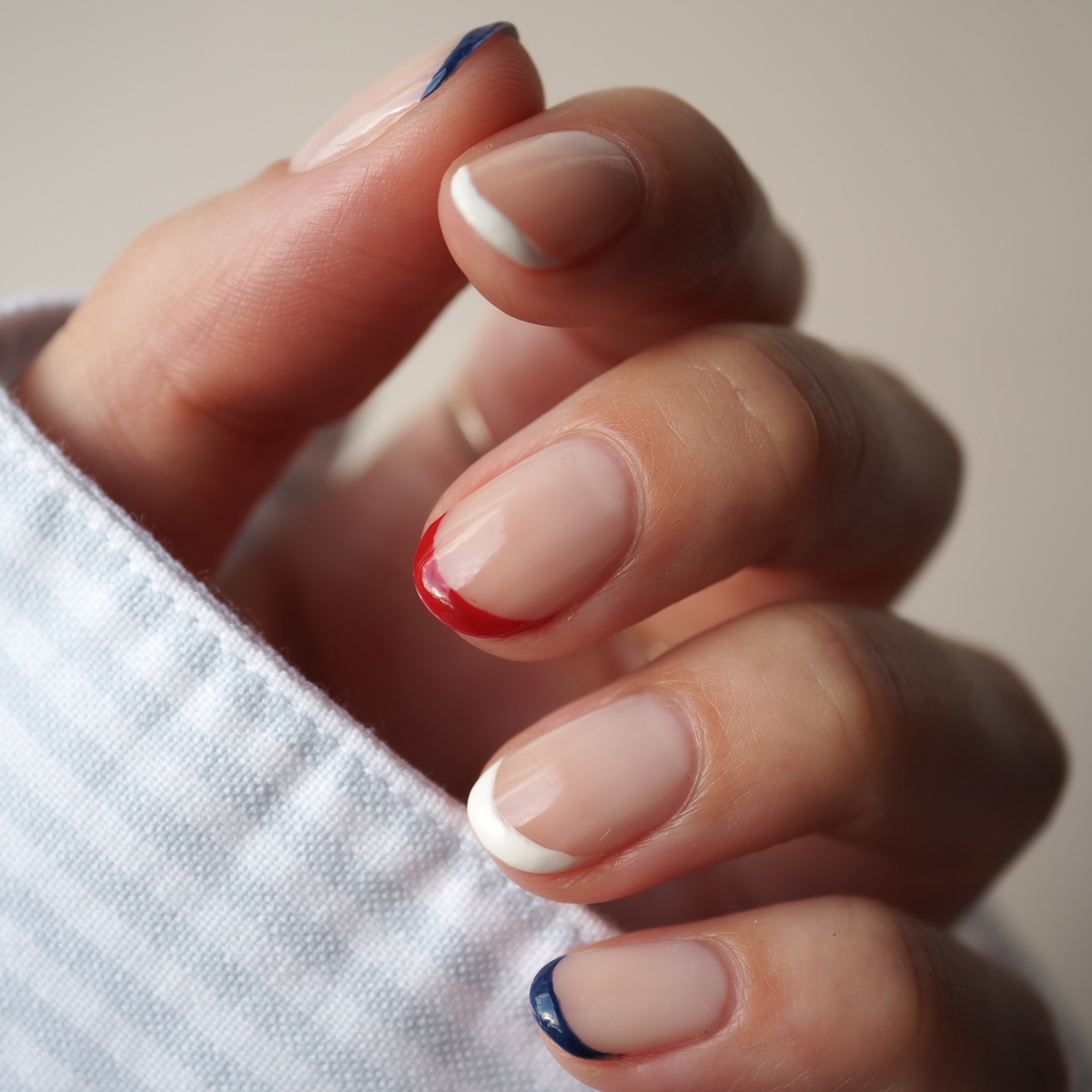 A hand with a neutral manicure using the Manucurist Red, White Blue Trio—blue on pinky and thumb, red on middle, white on index and ring—paired with a striped sleeve and matching bold red, white, and blue eyeshadow.