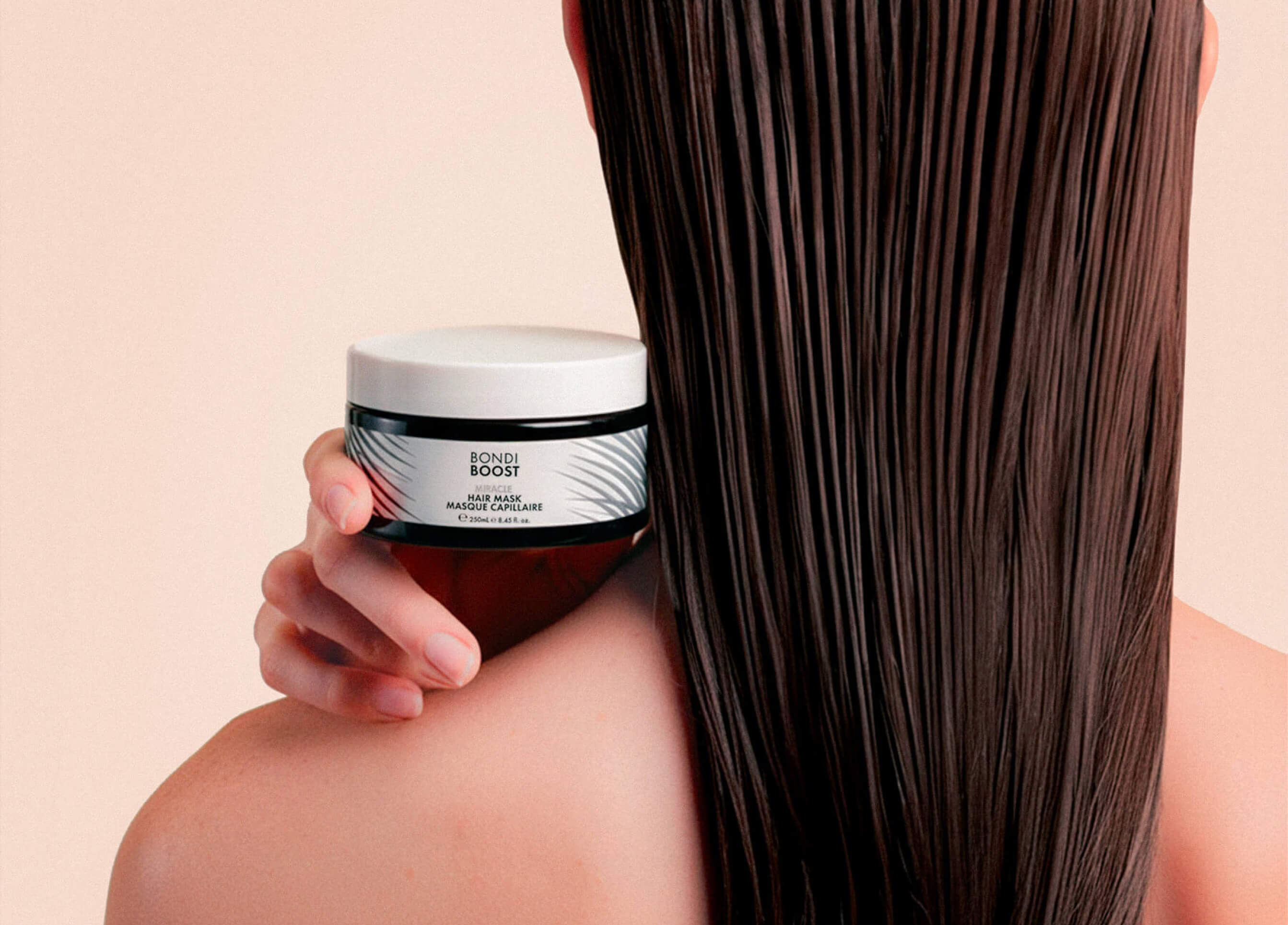 A person with straight, wet, brown hair holds a jar of Bondi Boost hair mask against their bare shoulder, displaying the product label.